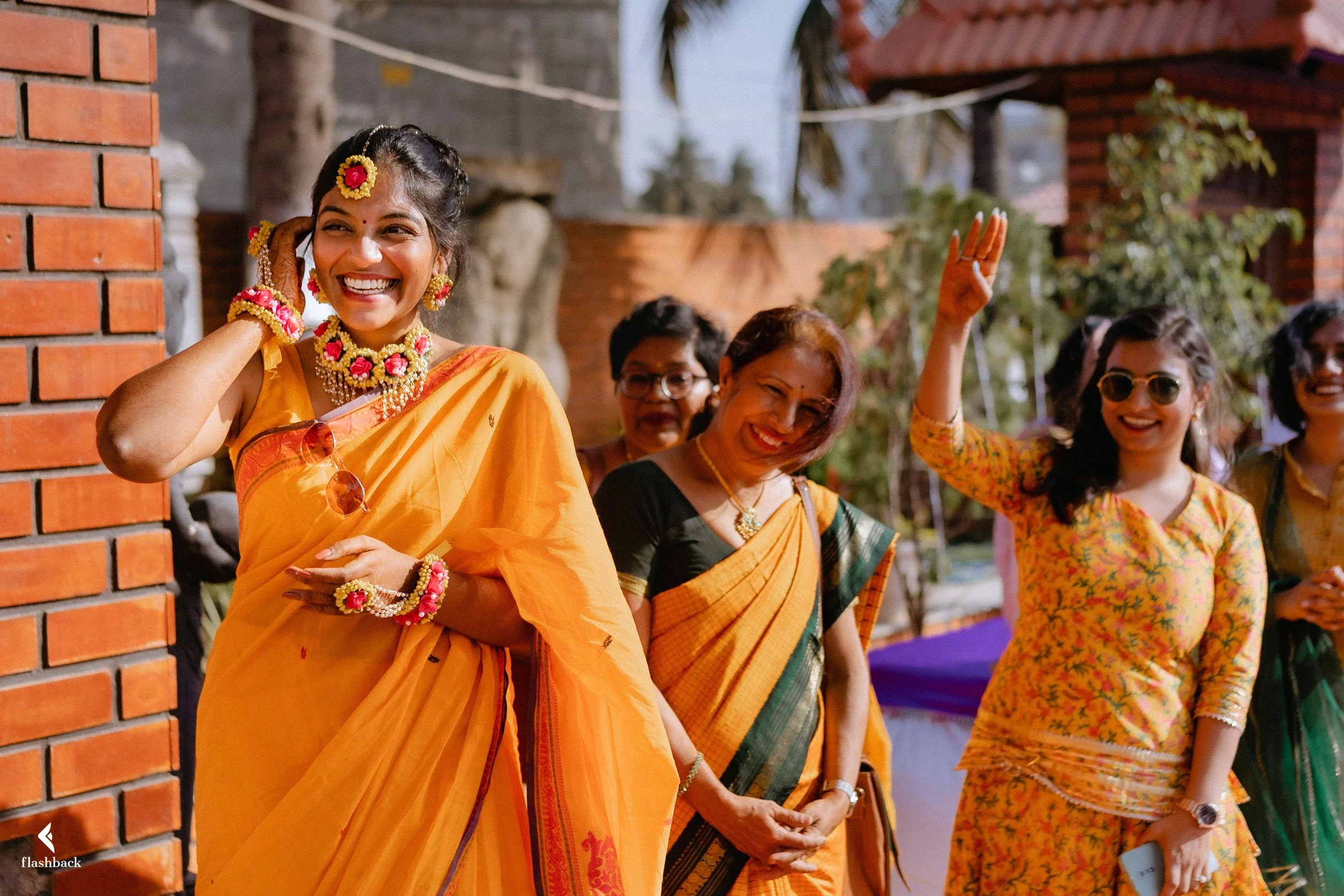 A group of women smiling and laughing, dressed in traditional Indian sarees and jewelry, gathered outdoors during daytime.