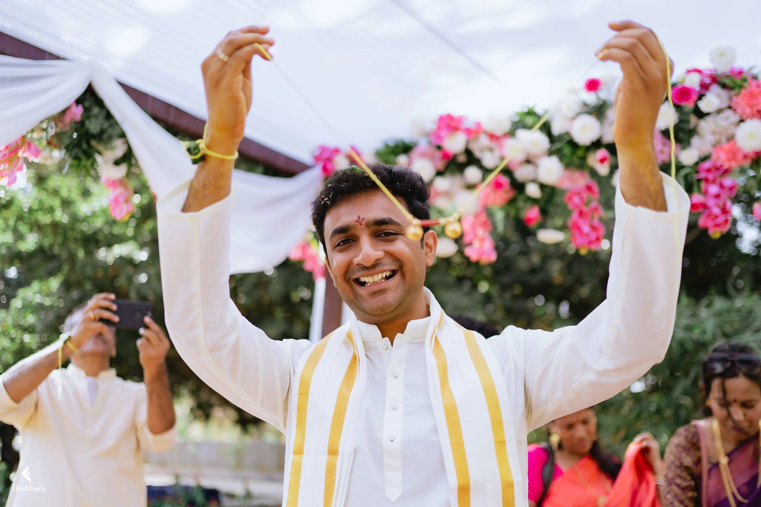 Happy man in traditional white Indian attire holding a gold ornament during a celebration outdoors with colorful flowers and people in traditional clothing in the background.
