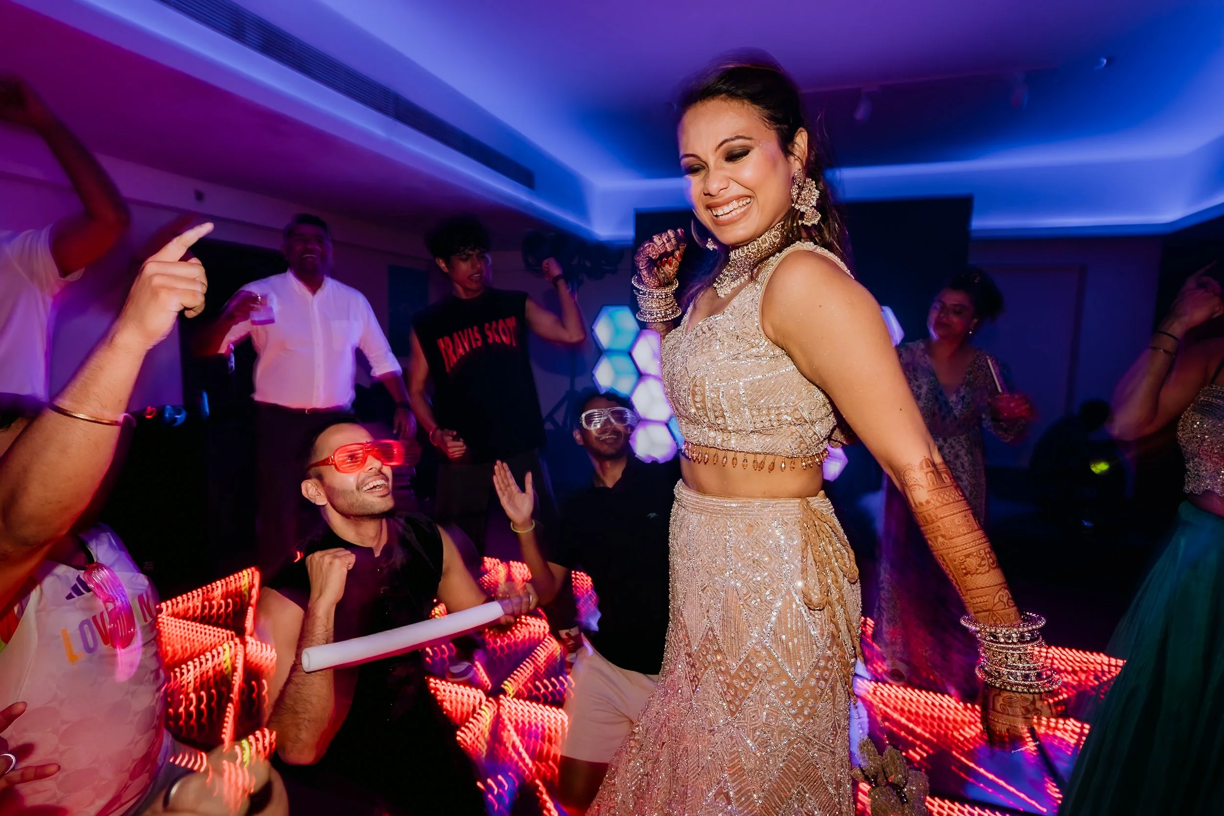 A woman in traditional Indian attire dancing at a lively celebration with friends. The room is decorated with blue lighting, and people around her are enjoying and dancing.