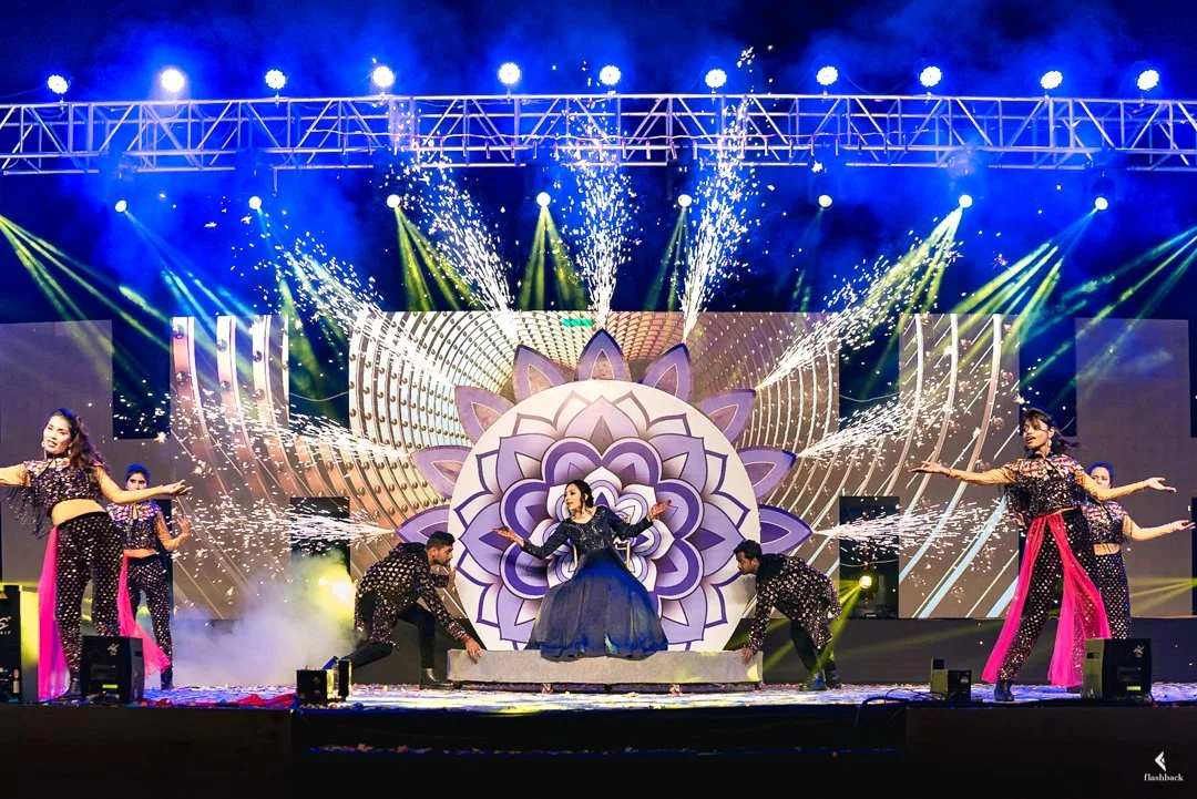 Stage with performers in colorful costumes, a large central floral design, lighting effects, and fireworks.