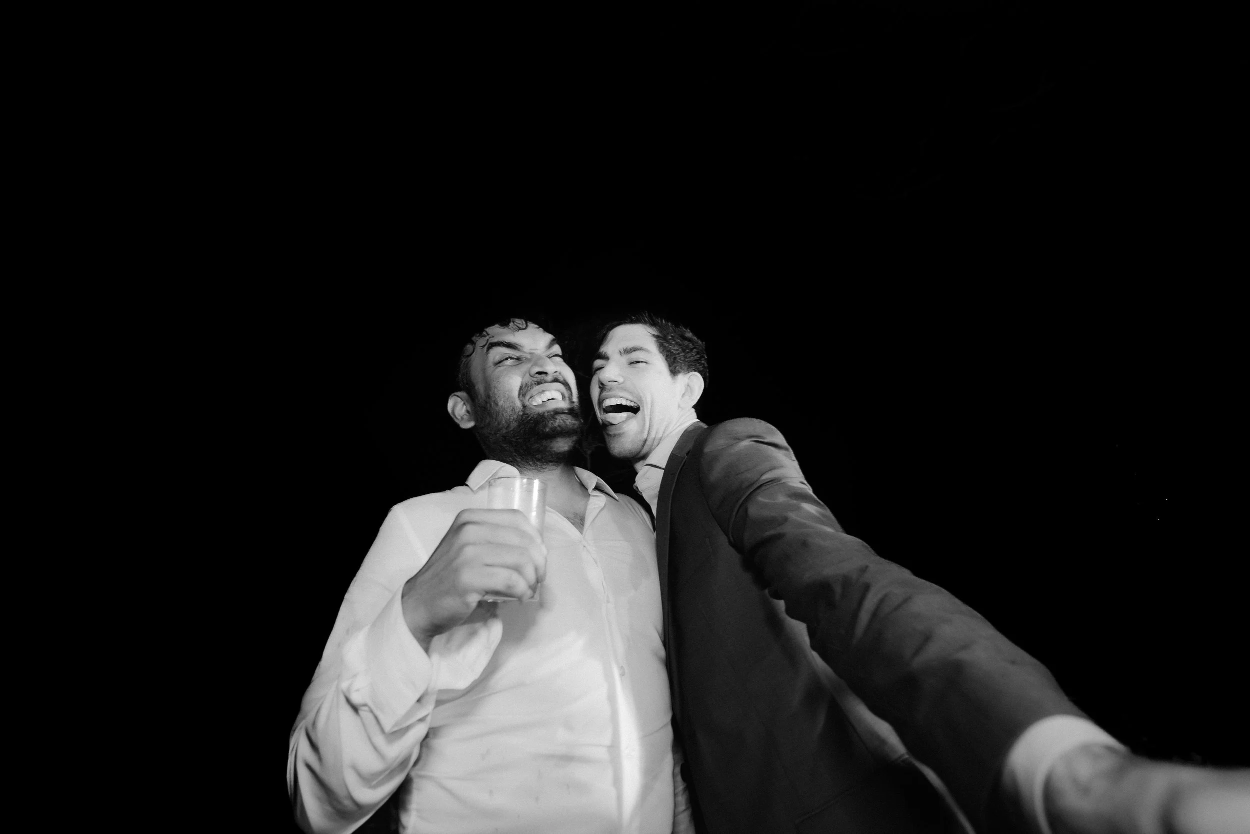Two men are smiling and laughing close together against a black background. One man holds a drink, and they appear to be celebrating.