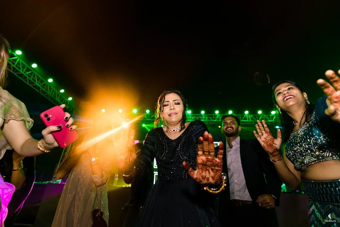 People dancing and celebrating at a lively event with colorful lights and a stage in the background.