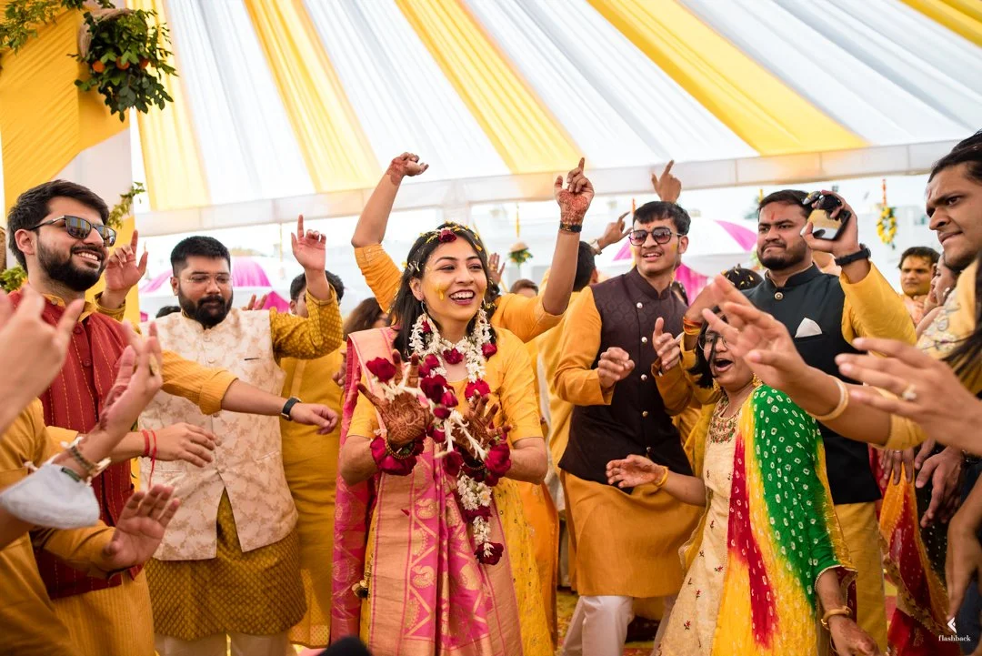 People celebrating at a traditional Indian wedding, dancing and smiling under a decorated canopy with yellow and white fabric.