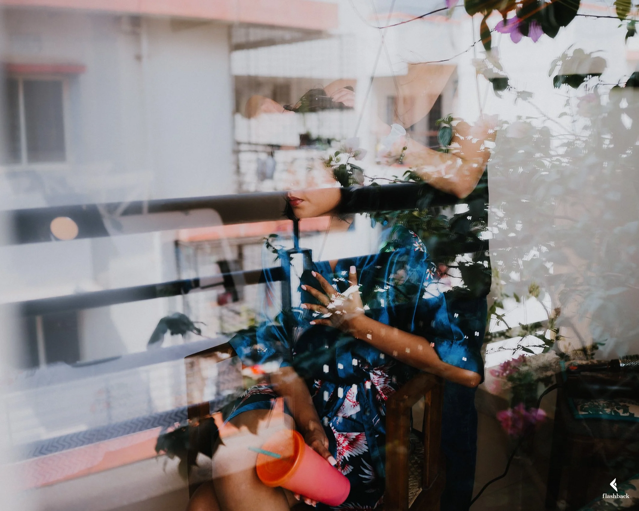 Woman sitting by a window, looking at her phone, with reflections of buildings and plants on the glass.