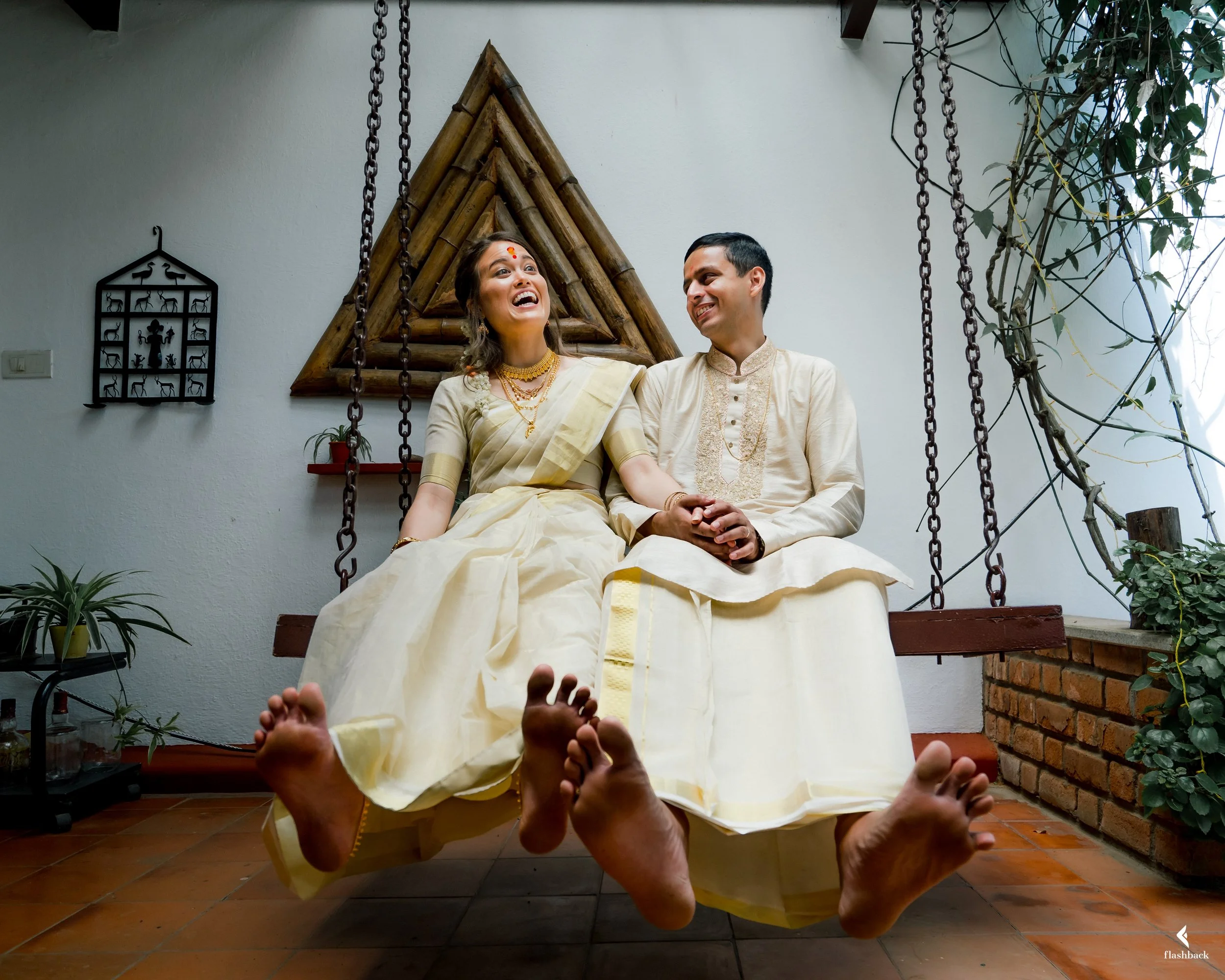 A couple in traditional Indian attire sitting on a swing inside a house, holding hands and smiling at each other.