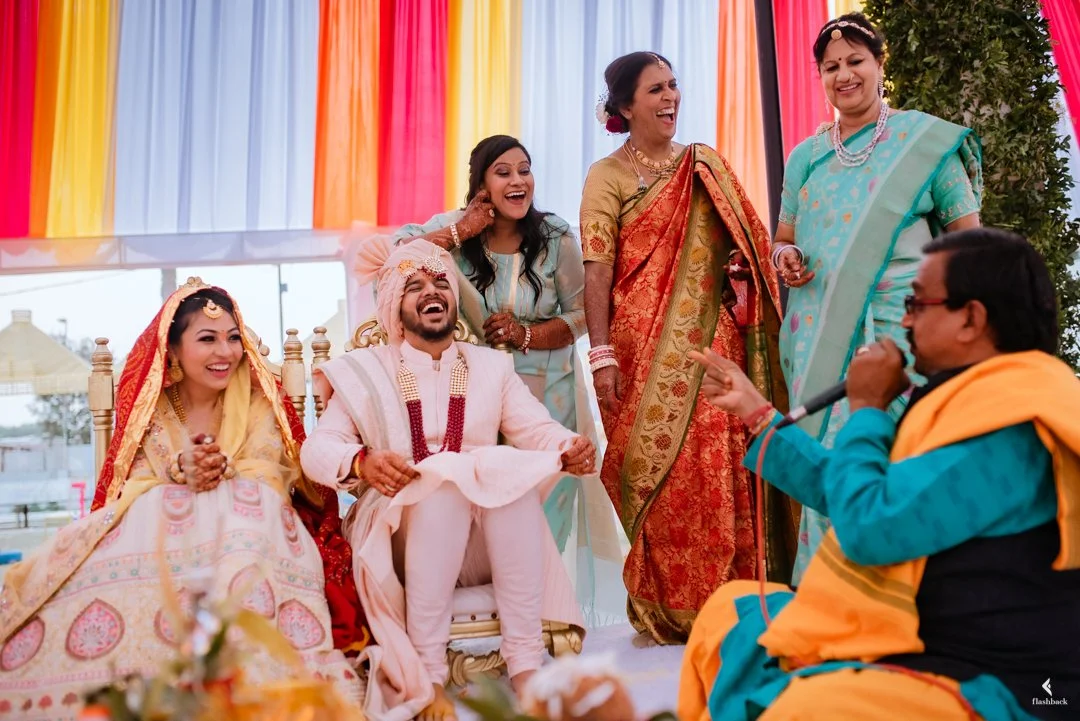 A traditional Indian wedding ceremony with the bride and groom sitting together, surrounded by family members. The bride wears a wedding saree and jewelry, while the groom is in a cream-colored sherwani. The officiant, dressed in bright colors, speak