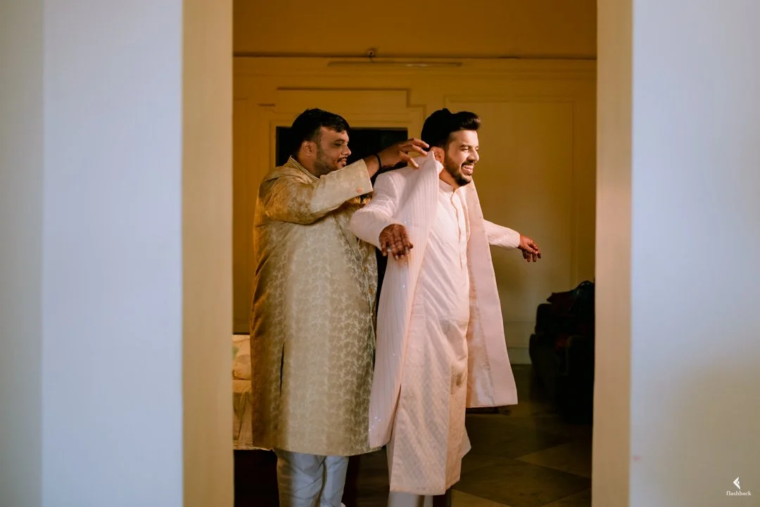 Two men dressed in traditional Indian attire smiling and helping each other get ready in a hotel room, possibly for a wedding.