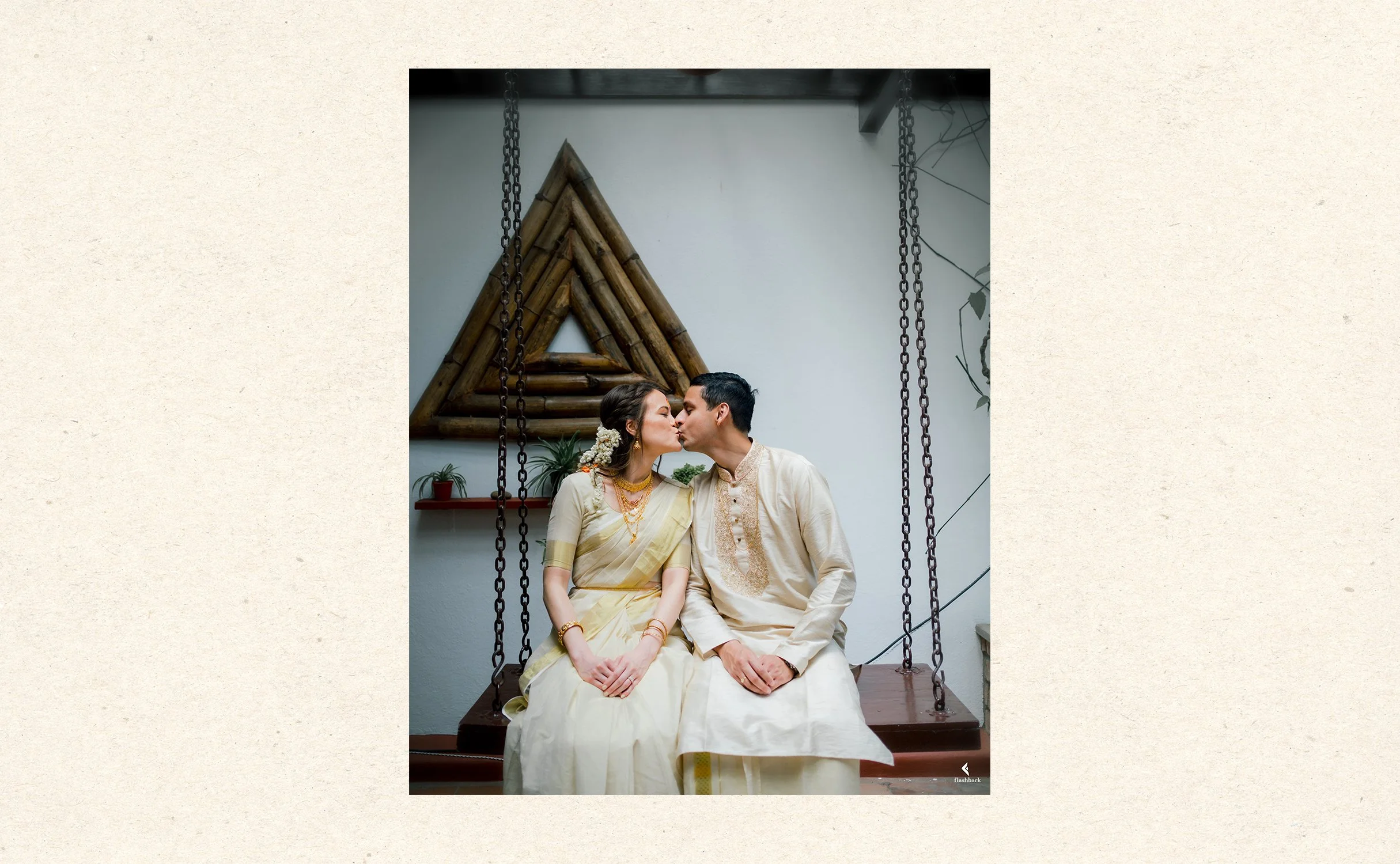 A couple dressed in traditional Indian attire sitting on a swing, about to kiss, with a decorative wooden triangle and potted plants in the background.
