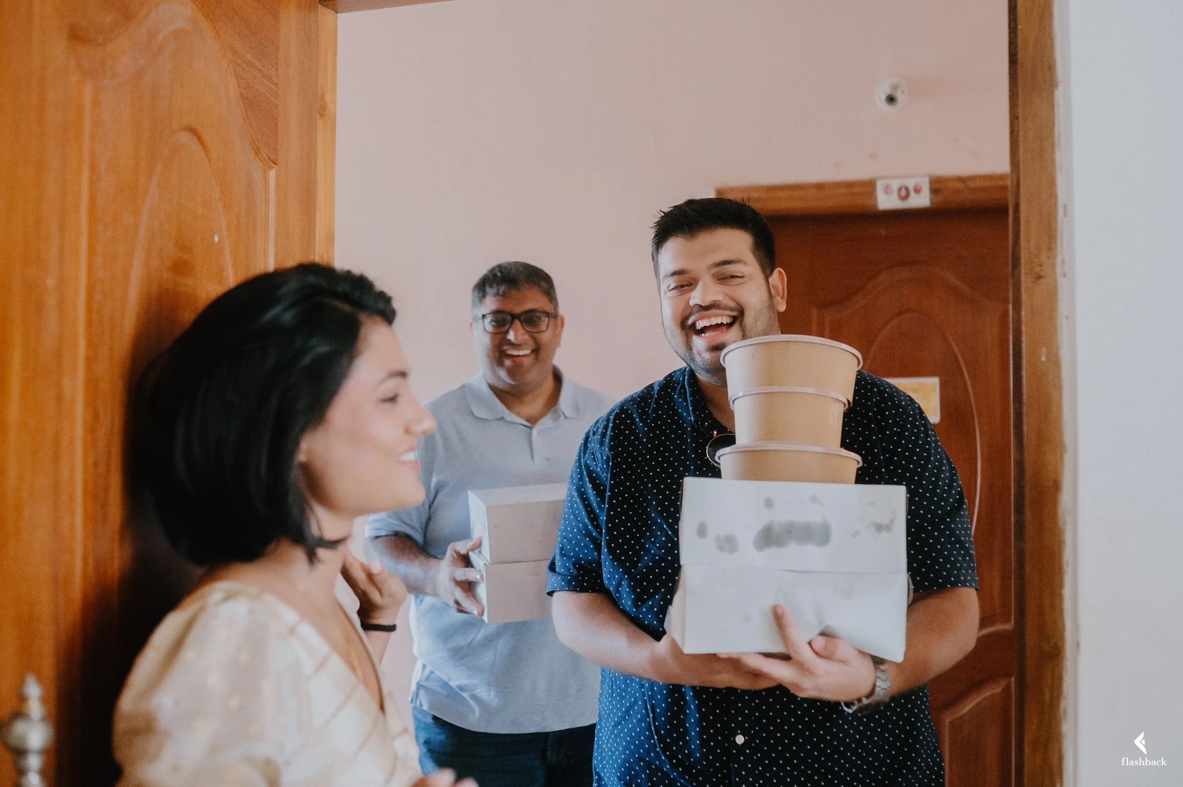 Three people smiling and holding boxes and plant pots in an indoor setting with wooden doors.