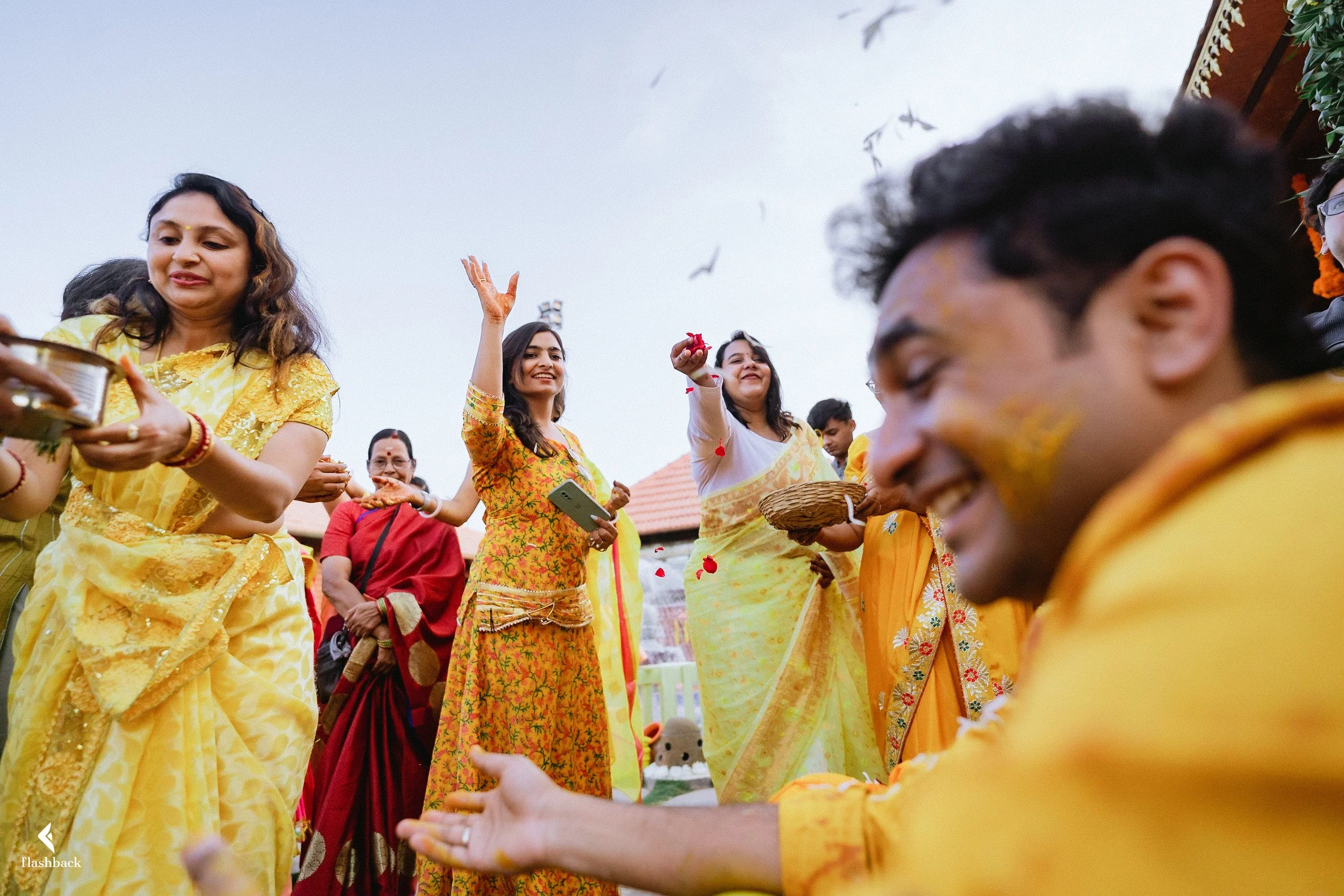 People celebrating a traditional Indian festival outdoors, with women and men dressed in colorful sarees and kurtas, some throwing flower petals and smiling, with a clear sky and rooftops in the background.