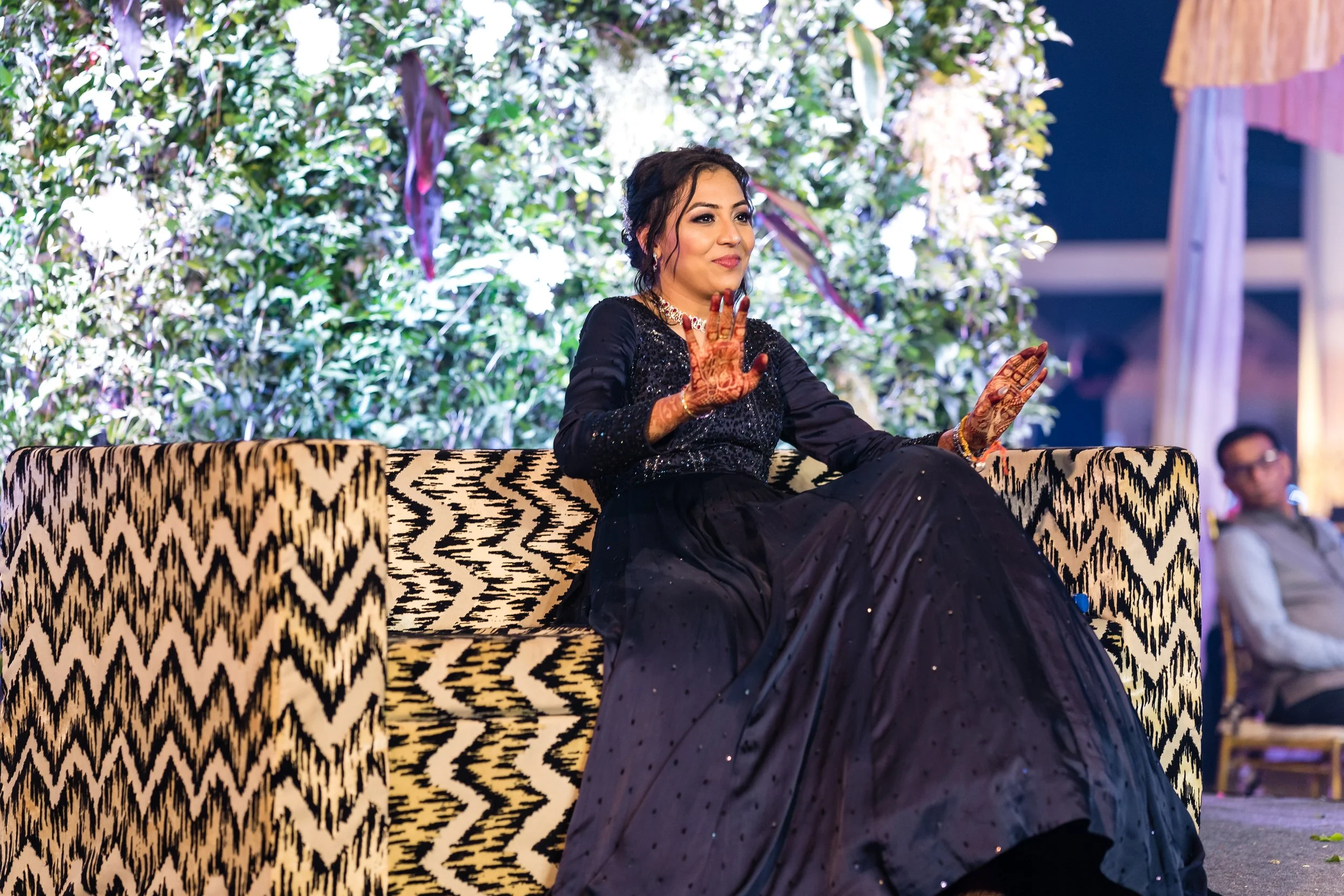 A woman dressed in a black saree with glittery details, sitting on a patterned sofa, displaying hand gestures, at a decorated event with greenery and lighting in the background.