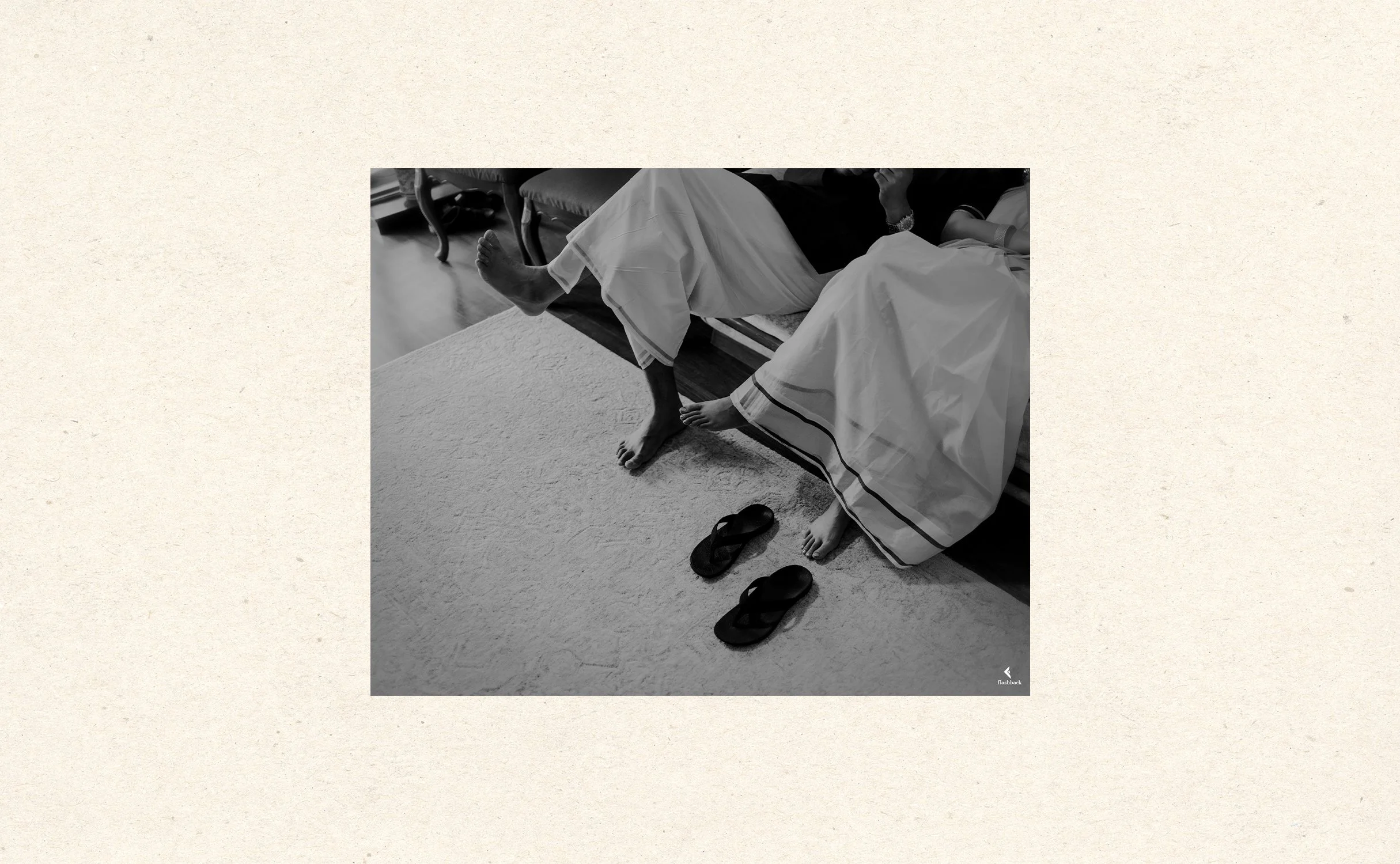 Two people sitting on a bed with their feet hanging off, wearing traditional white garments. Two pairs of black sandals are on the floor next to the bed.