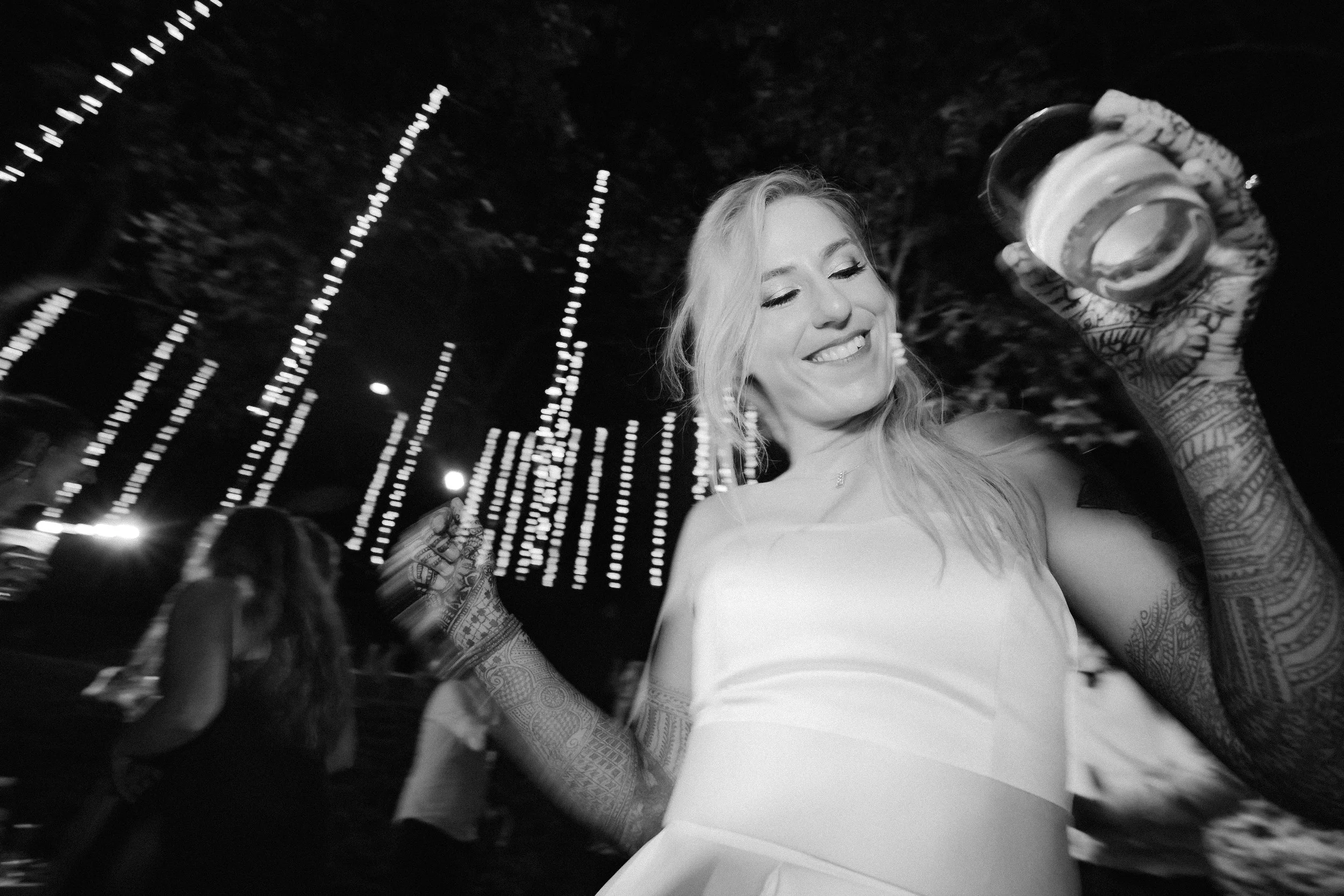 A smiling woman with tattoos on her arms holding a glass of wine at night, with decorative string lights in the background.