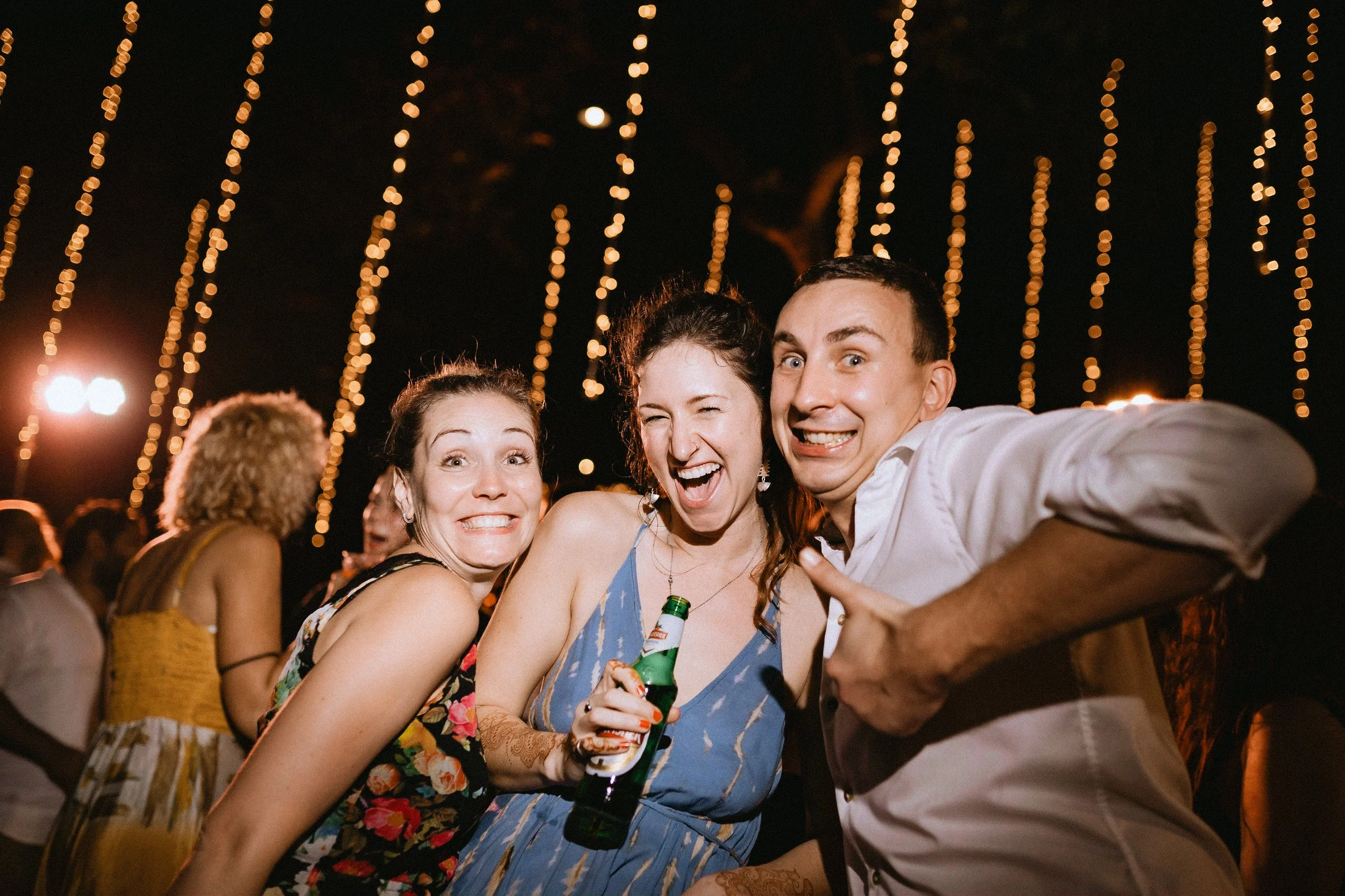 Three friends smiling and posing for a photo at an outdoor night event with string lights overhead. The woman in the middle is holding a beer bottle, and all are making playful expressions.