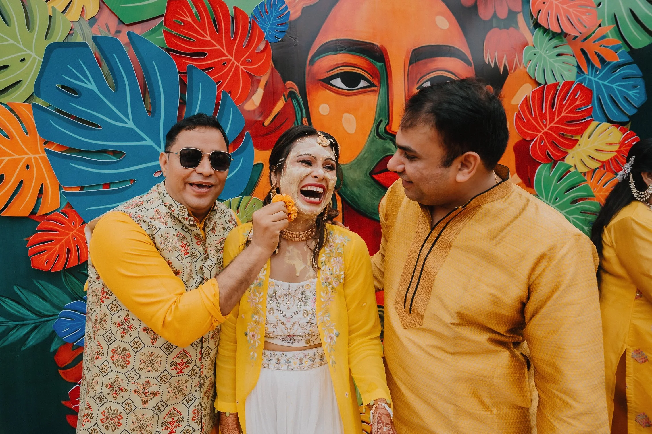 People celebrating at a colorful event, with a backdrop of vibrant tropical leaves and an artistic face portrait. The woman in the center is smiling and applying turmeric or a similar substance on her face, indicating a festive or cultural celebratio
