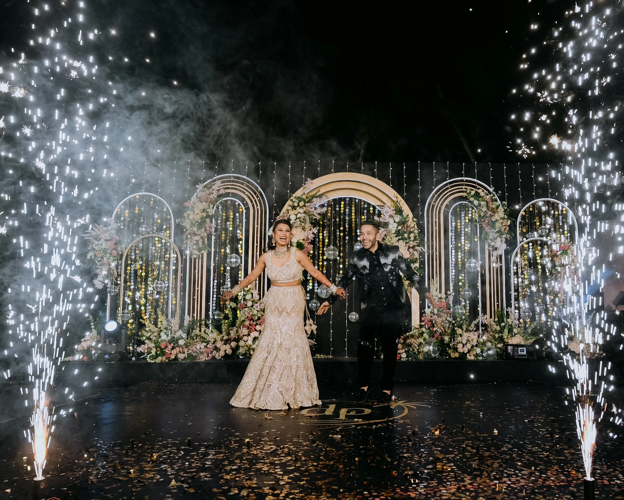 A newlywed couple celebrating at their wedding reception, standing in front of a decorated backdrop with flowers and arches, fireworks and sparklers surrounding them, and a dark night sky overhead.