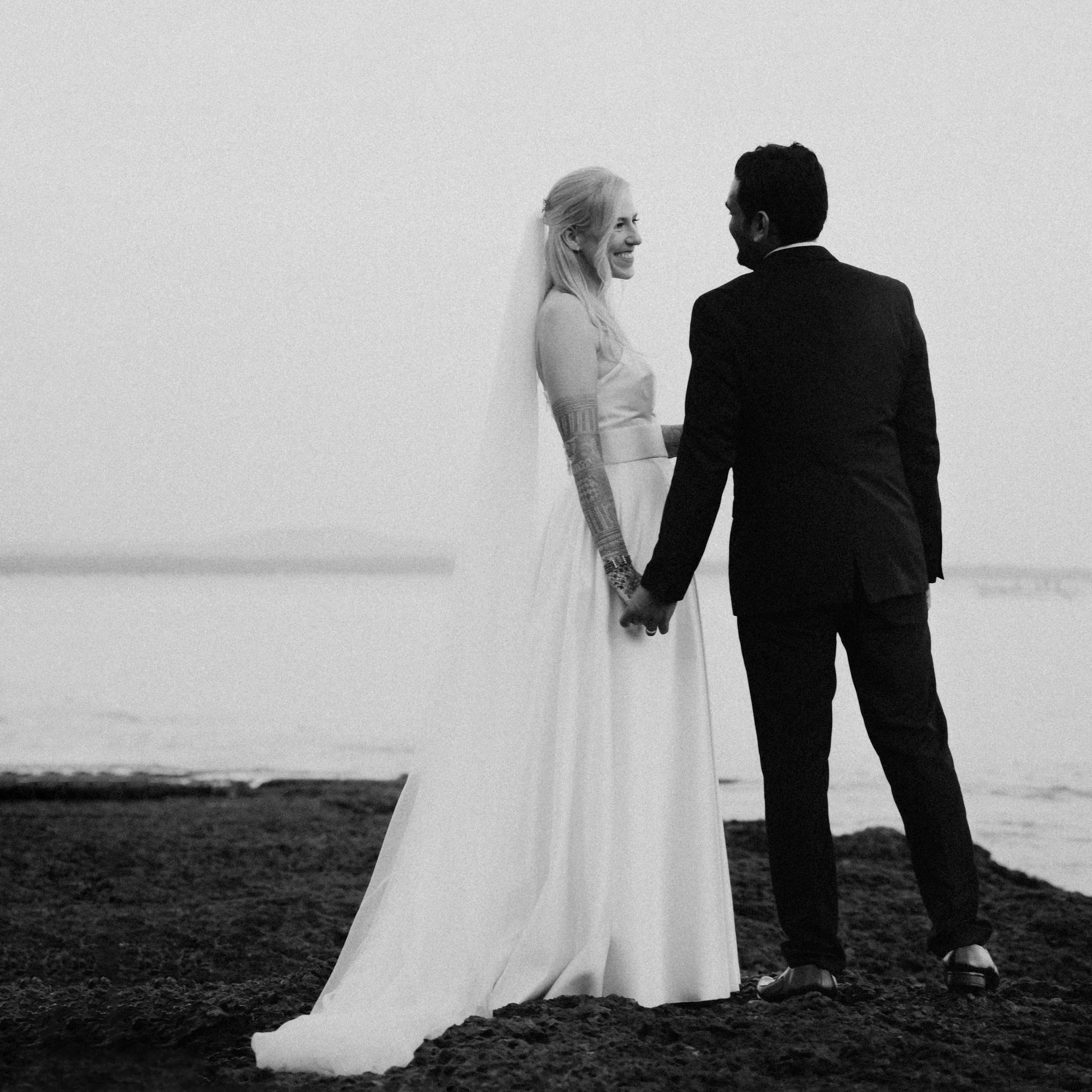 A couple holding hands on the beach, the woman wearing a wedding dress and veil, smiling at each other.
