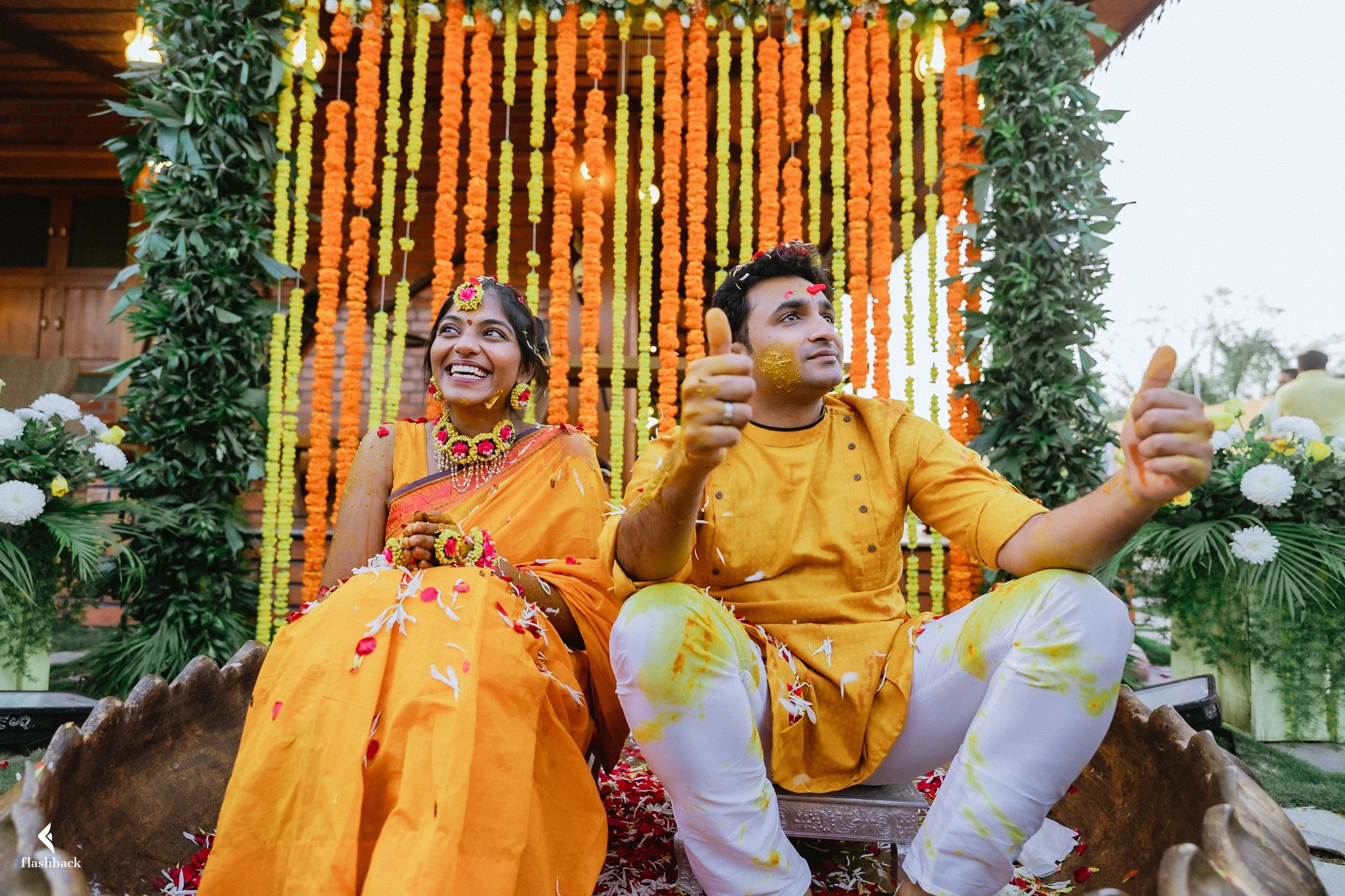 A happy couple sitting on a decorated wooden platform during a traditional Indian wedding. The woman is smiling and wearing a yellow sari with floral jewelry, while the man is giving a thumbs-up and wearing a yellow kurta with white pants. The backdr