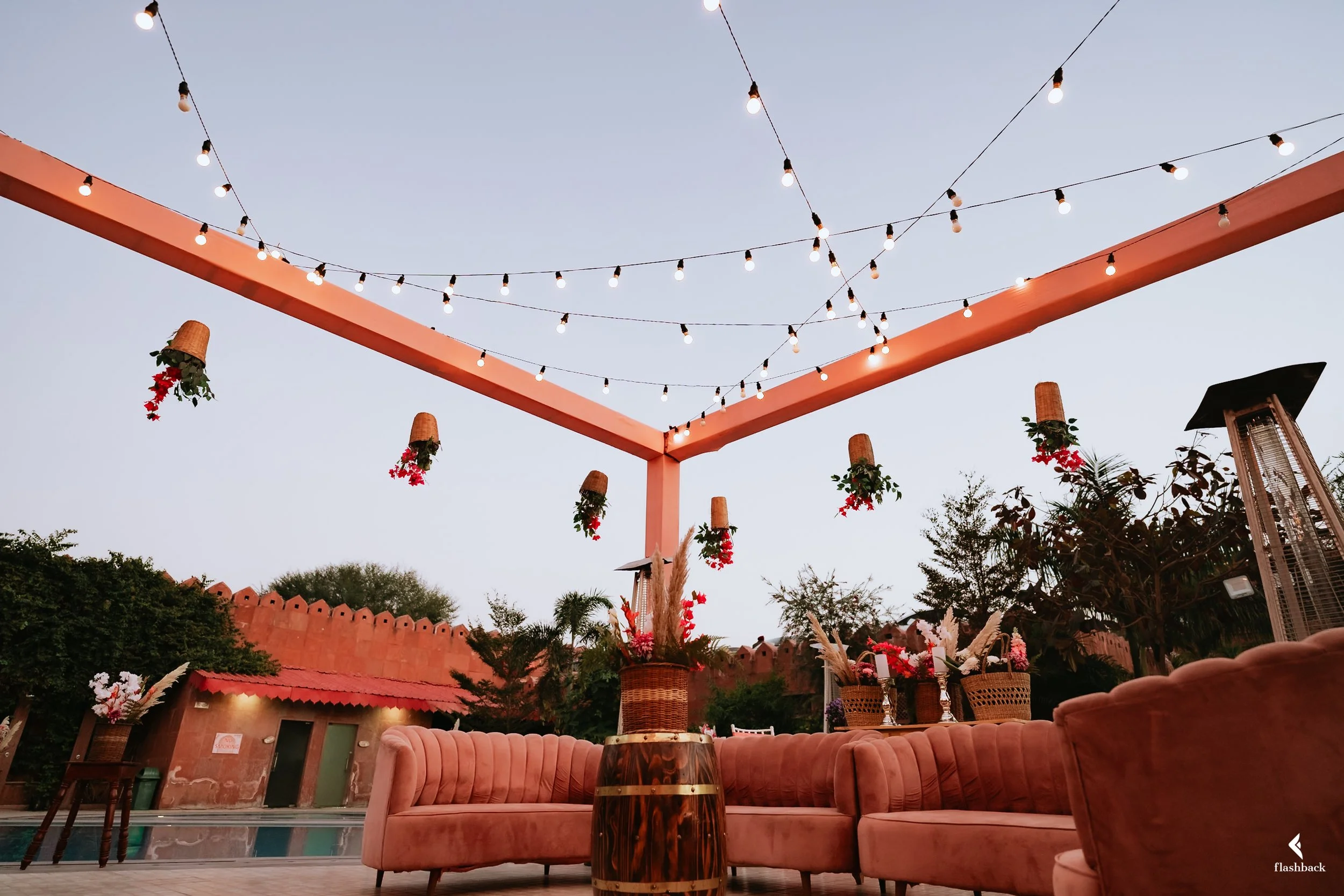 An outdoor patio decorated with string lights hanging from a pink structure, flower baskets, pink velvet sofas, and a central vase with flowers, set against a clear evening sky and a wall with small arches.