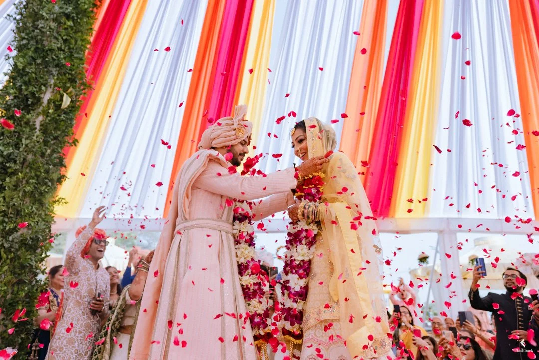 A traditional wedding ceremony with a bride and groom dressed in pink and gold, exchanging garlands under a decorated canopy, with guests throwing flower petals.