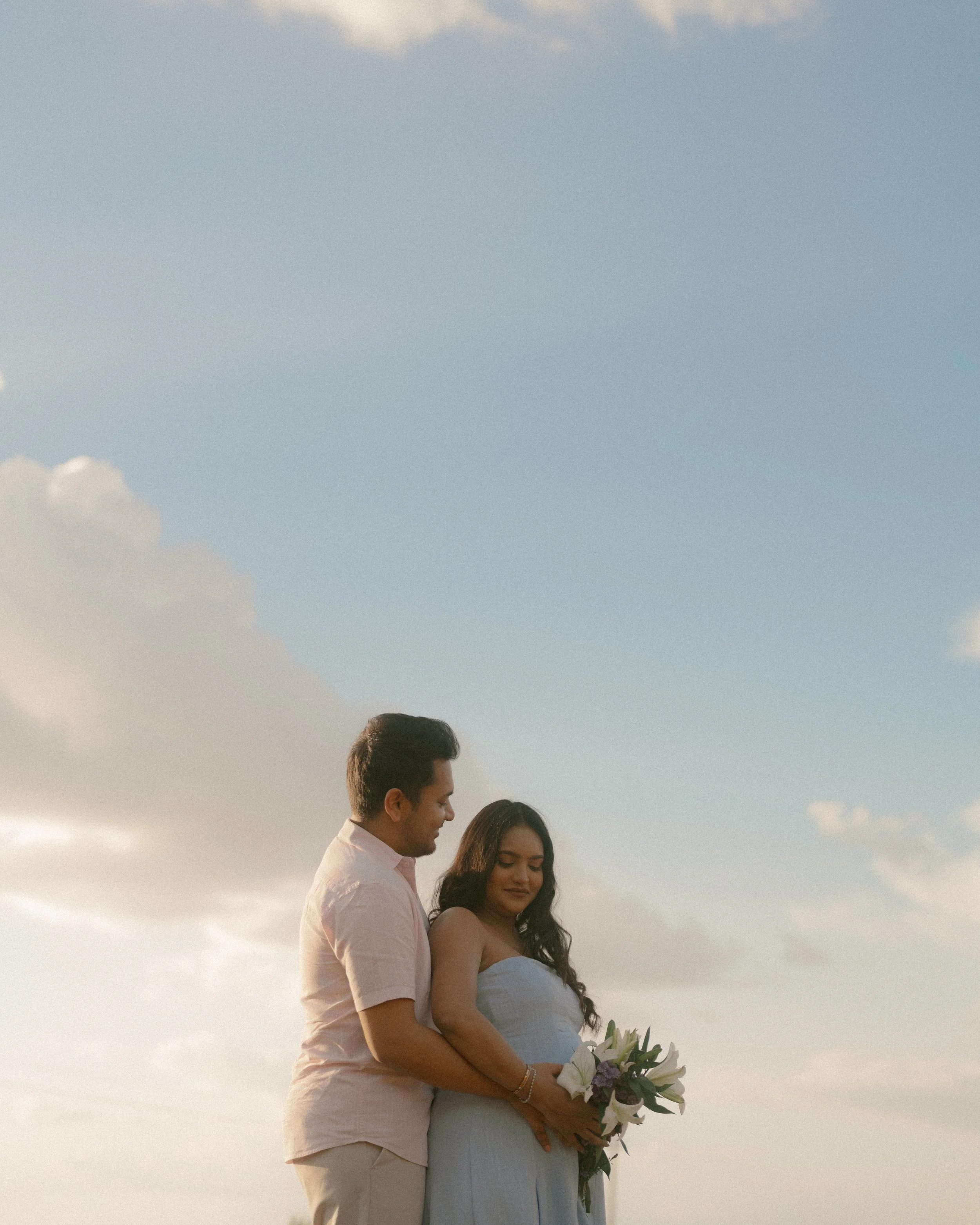 Couple embracing under a wide blue sky, holding a bouquet of lilies