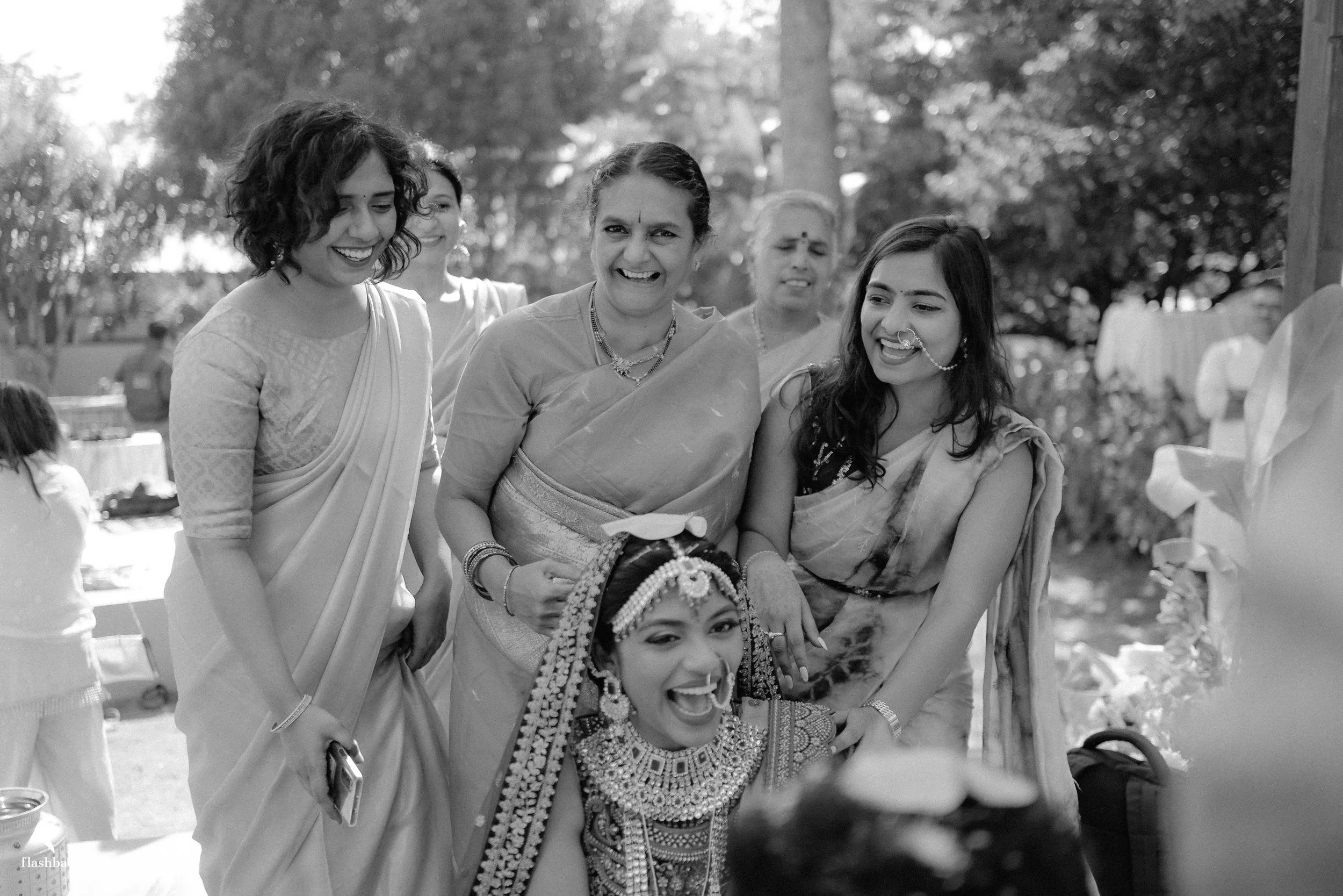 Group of women in traditional Indian attire smiling outdoors, with one woman dressed in bridal jewelry and attire, during a celebration or wedding.