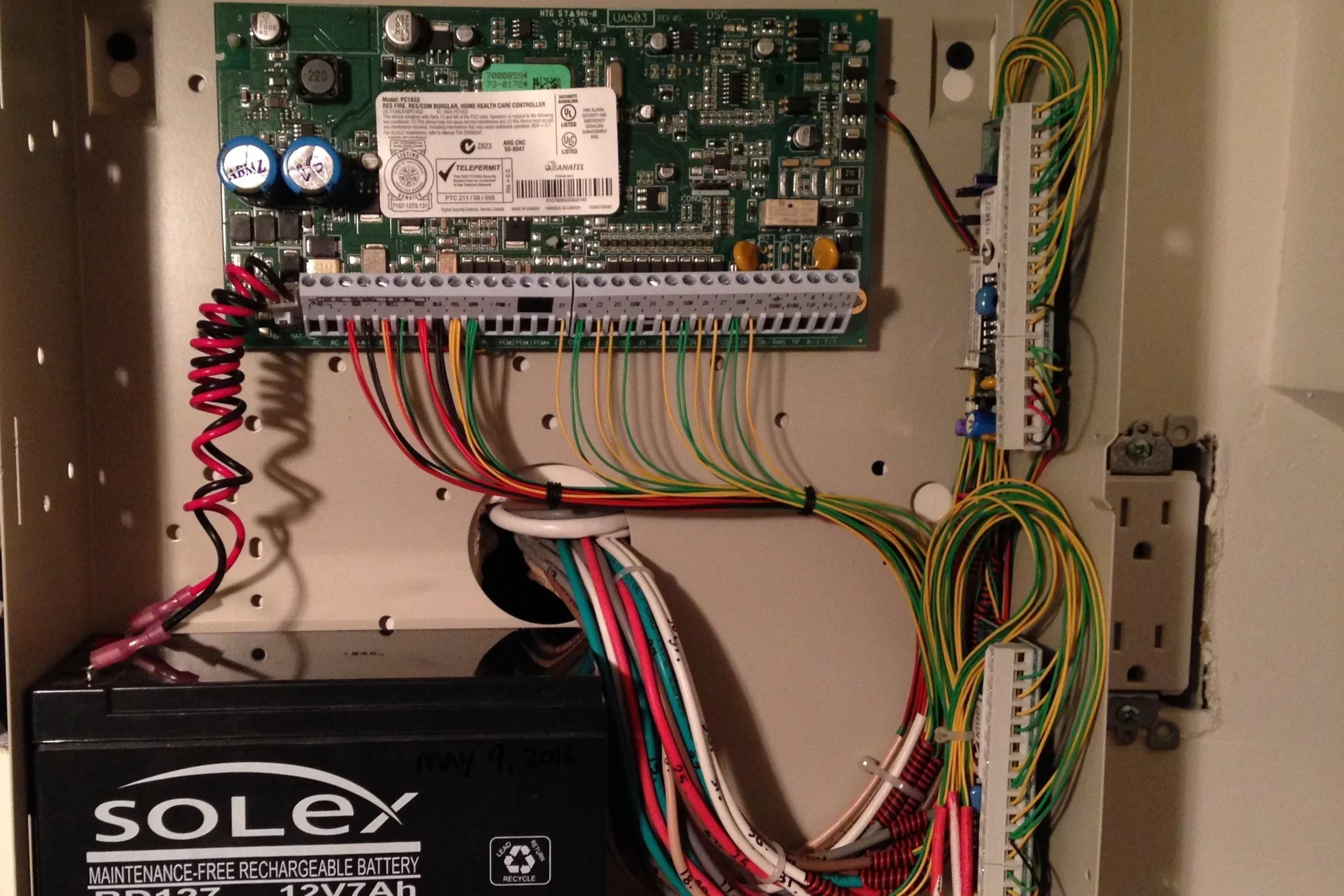 The interior of an electrical control panel with a circuit board, various colored wires, and a Solex rechargeable battery.