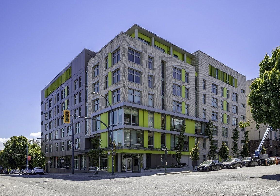 Modern multi-story building with gray brick facade and green accents, located on a city street with parked cars and trees.