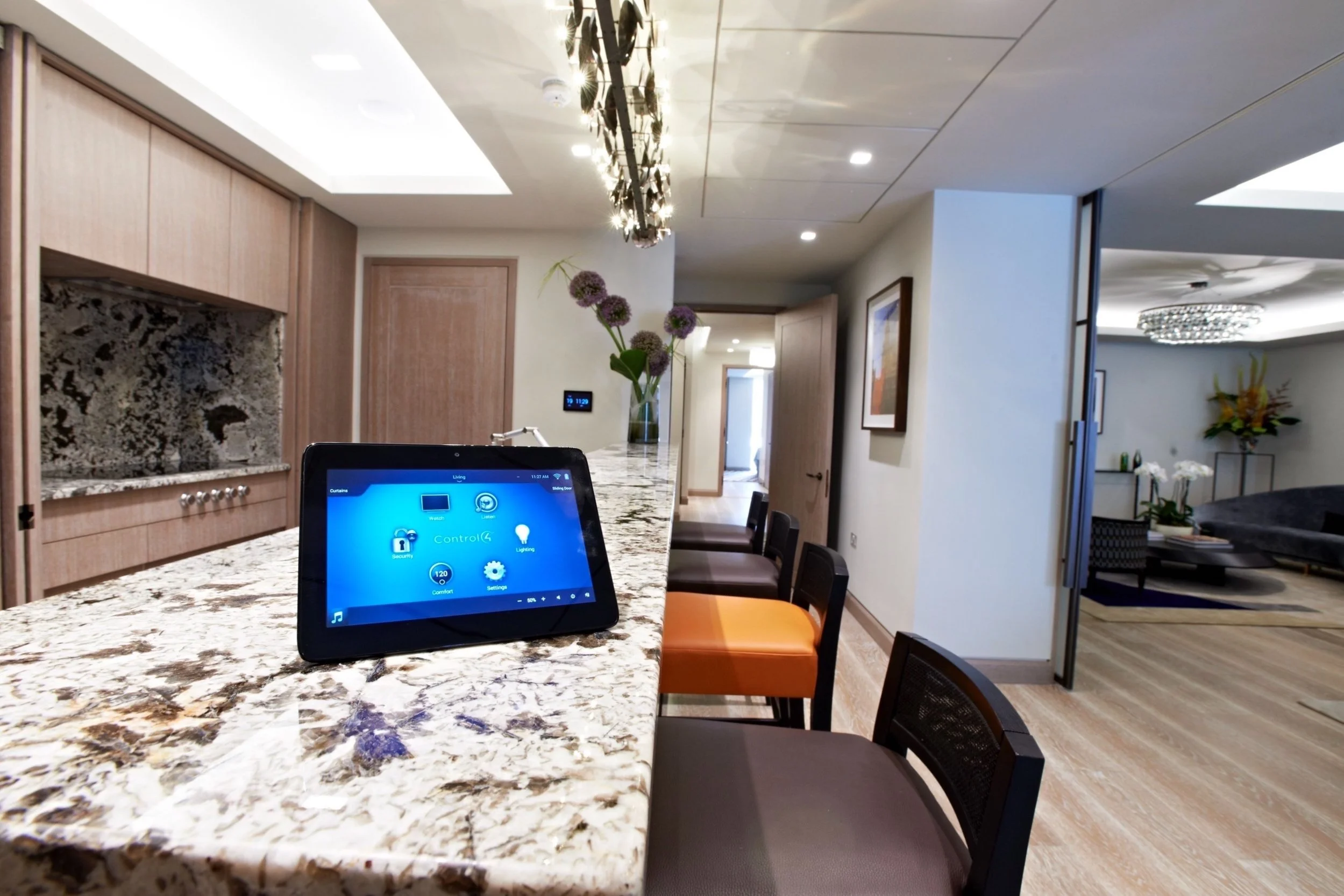 Modern living space with a granite kitchen island, a control tablet, and colorful chairs, leading to a lounge area with decorations and a chandelier.