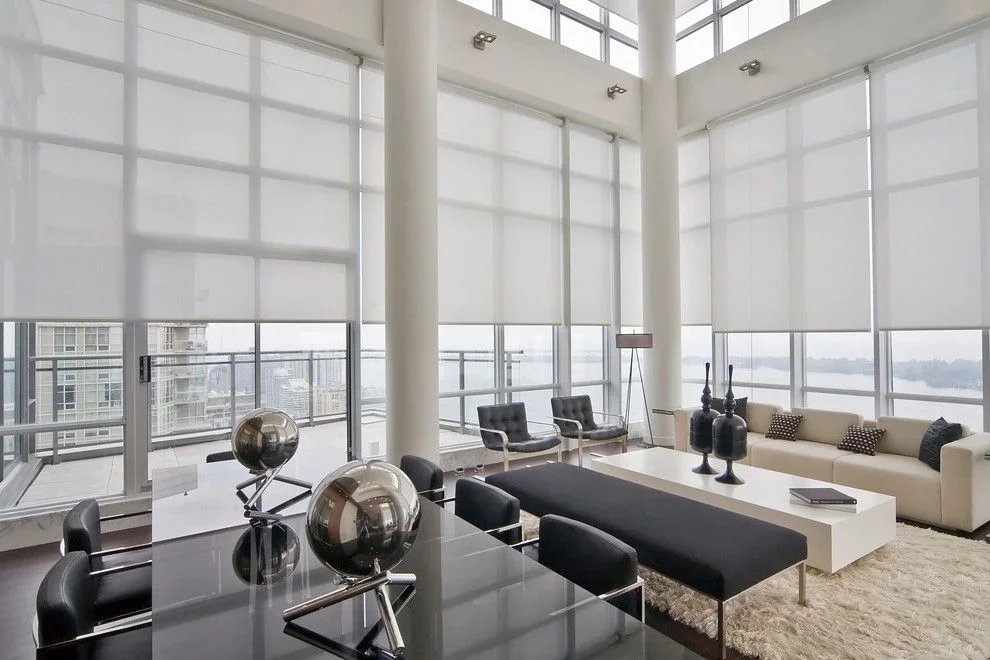Modern living room with large windows, white blinds, a cream sofa, black and gray chairs, a black and white coffee table, and city views