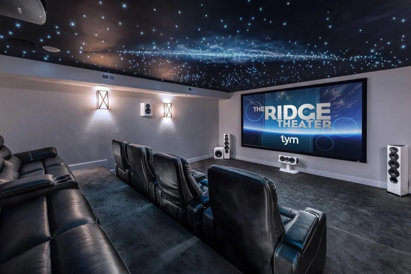 A home theater room with a large screen displaying 'The Ridge Theater'. The ceiling features a starry sky effect, and several black leather chairs are facing the screen. There is a gray carpet and white walls with wall lights.