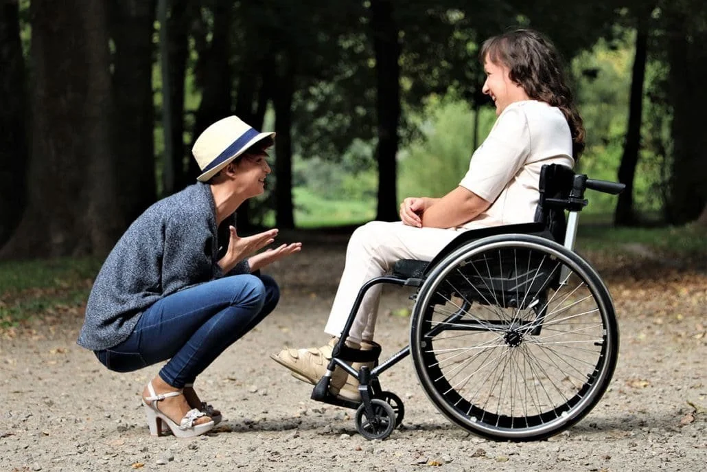 A woman with a hat and casual clothes is crouching and smiling at a woman sitting in a wheelchair, outdoors in a park with trees in the background.