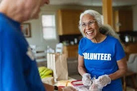 An elderly woman in a blue volunteer shirt smiling while interacting with a person in a kitchen or community space.