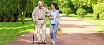 An elderly man with a cane walking and talking with a young woman carrying a bag in a park with green trees.