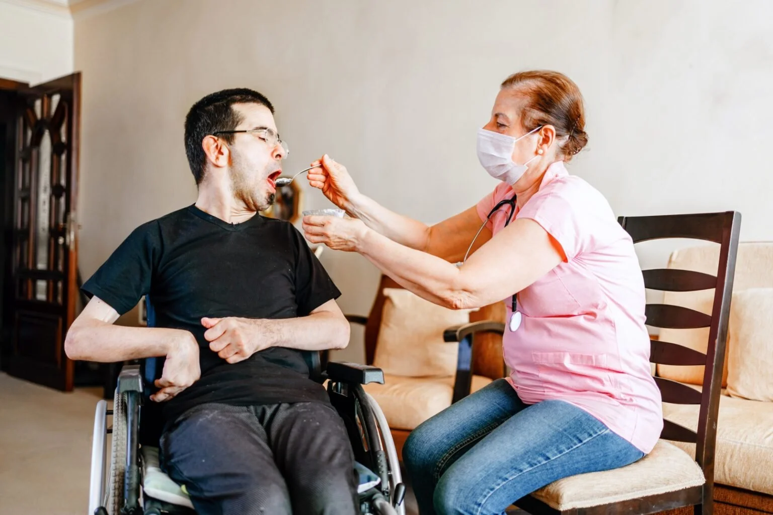 A nurse feeding a man in a wheelchair at home, wearing a face mask and pink scrubs.