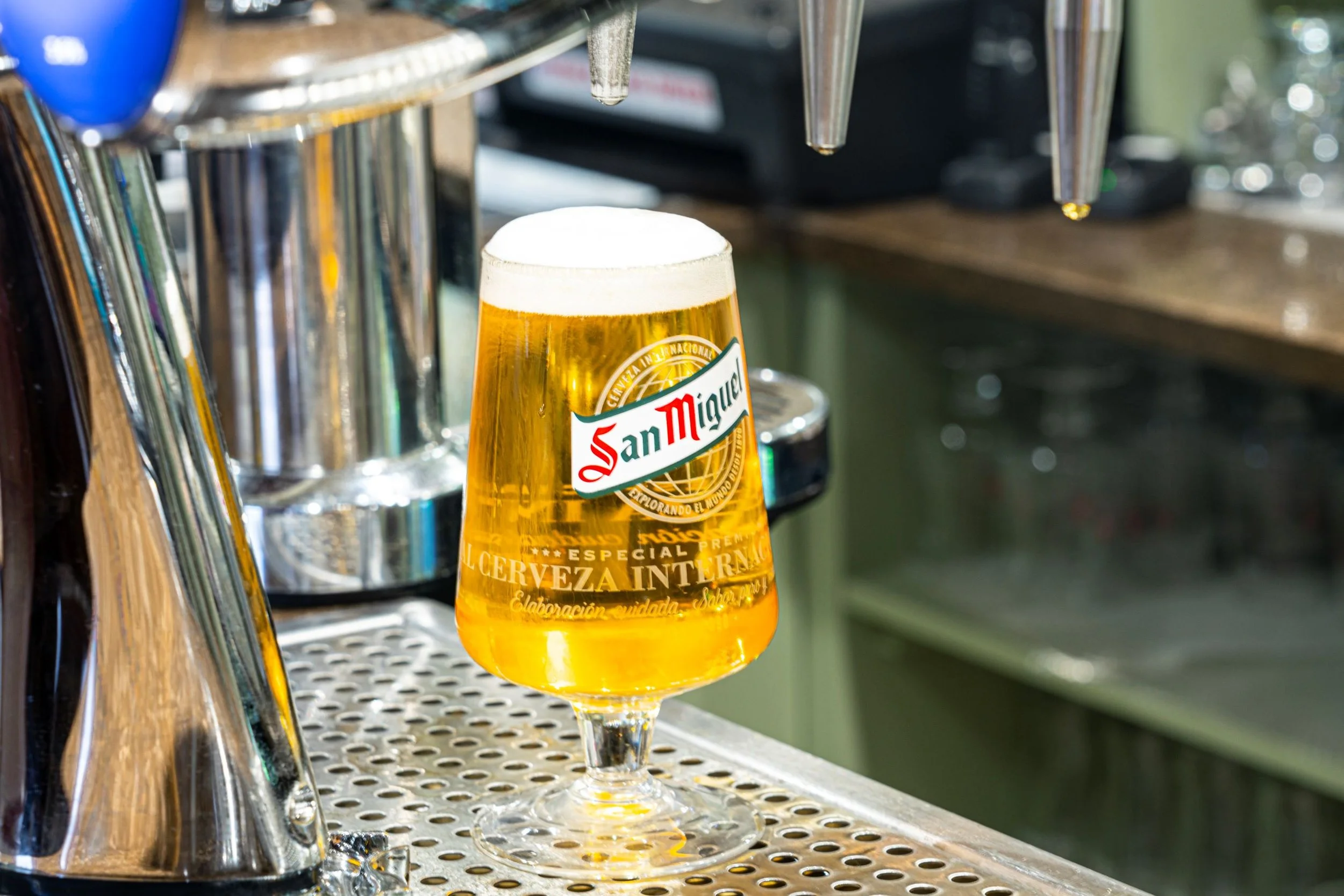 A glass of San Miguel beer on a bar counter next to beer taps.