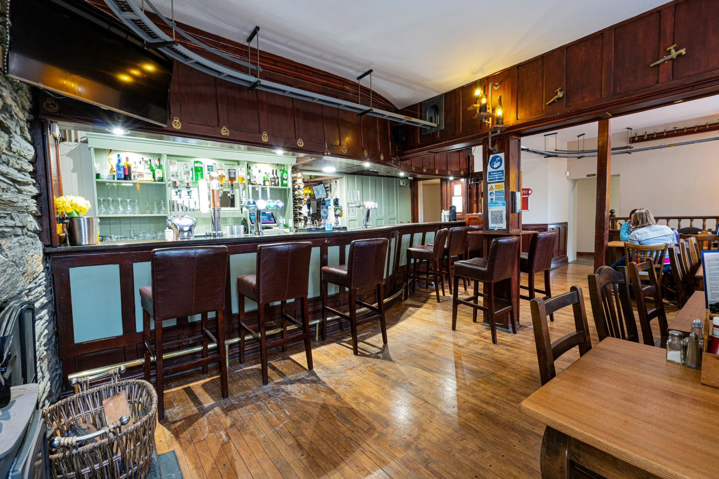 Interior of a rustic pub or restaurant with a bar counter, wooden chairs, and tables, two people sitting at one table in the background, and a TV mounted on the wall.