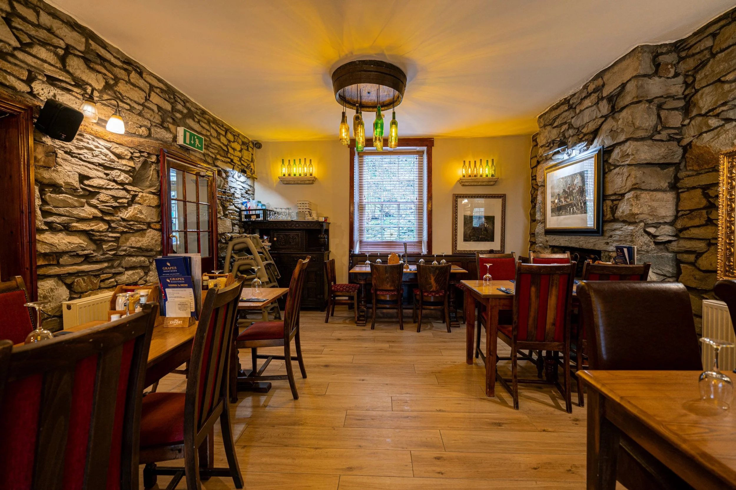 Interior of a cozy restaurant with wooden floors, stone walls, and wooden furniture. There are chairs and tables set for dining, some with glassware. A window with blinds is in the background, and decorative bottles are on shelves on the walls. The c
