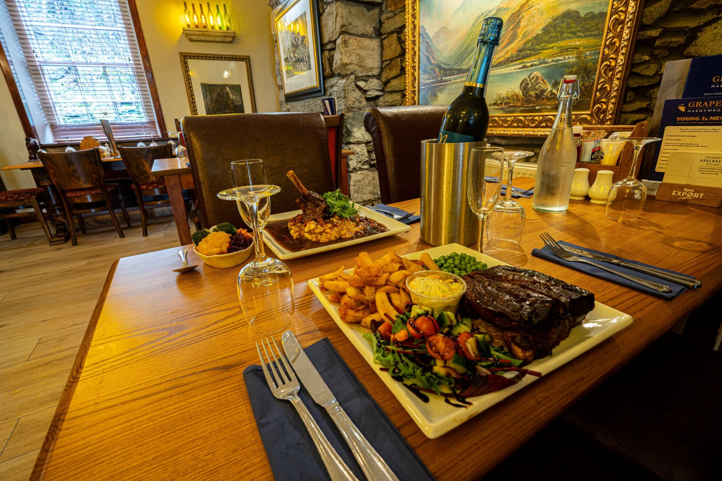 A wooden dining table set with a large plate of barbecue ribs, salad, french fries, and condiments, a smaller plate with a plated lamb chop dish, and several upside-down wine glasses, in a cozy restaurant with warm lighting and artwork on the walls.