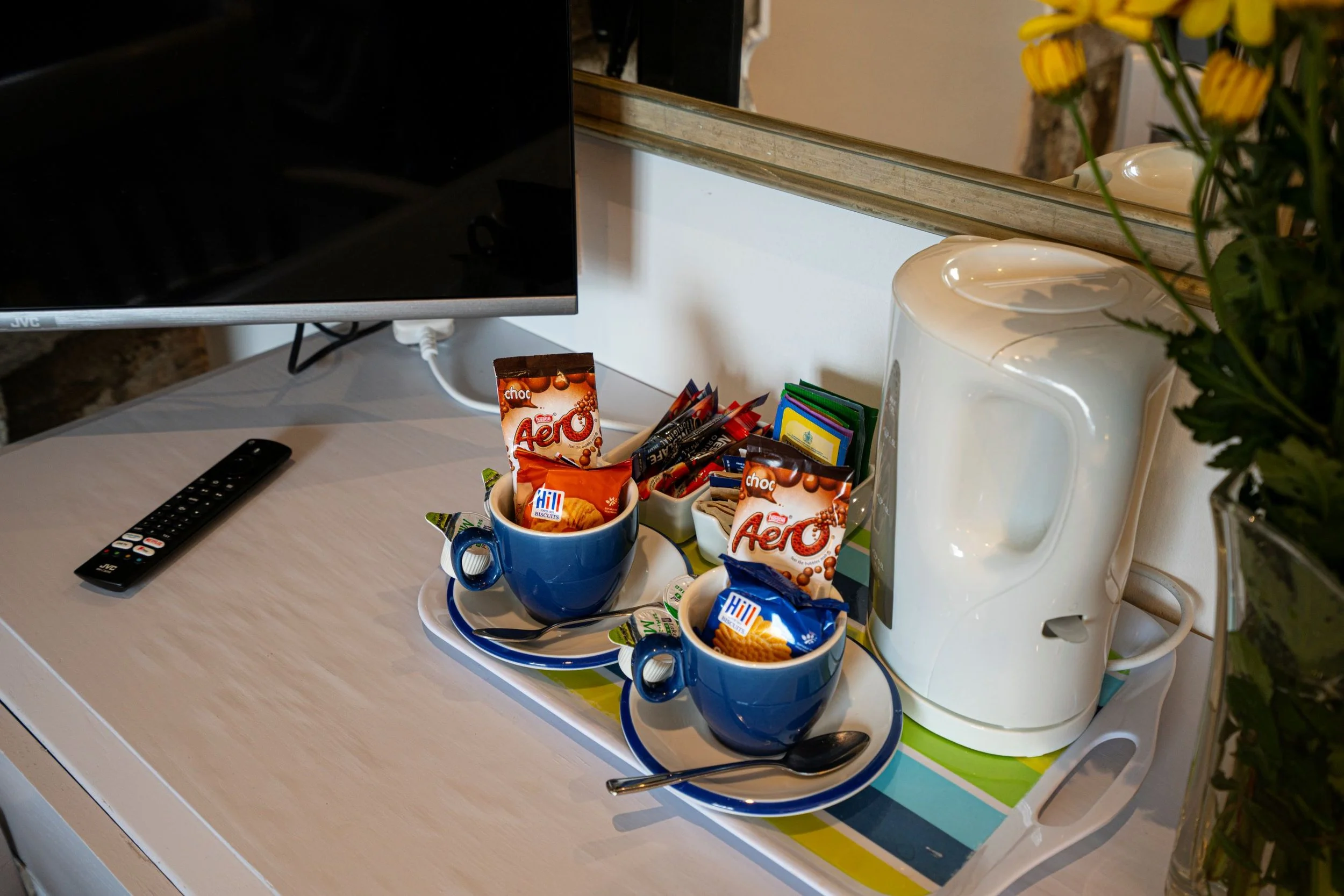 Tea or coffee setup with two blue cups filled with packets of Aero and Hill biscuits, a white electric kettle, a remote control, and a flower arrangement in the background on a white surface.