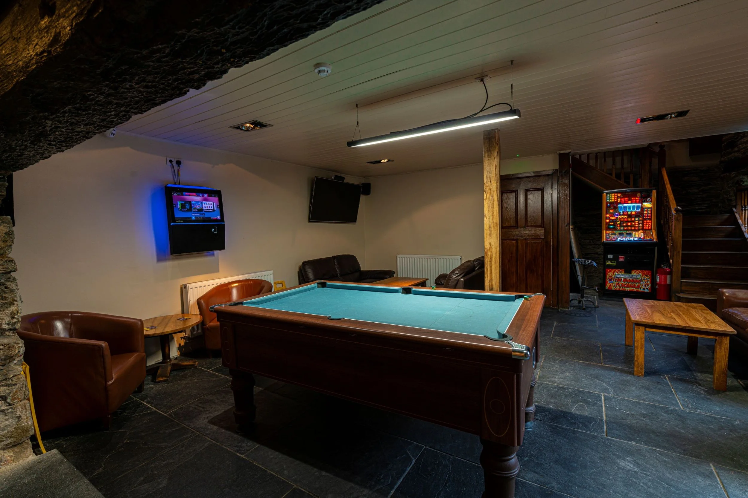 Indoor game room with a pool table in the center, surrounded by leather chairs, wall-mounted televisions, and a slot machine in the background, featuring dark stone flooring and wooden accents.