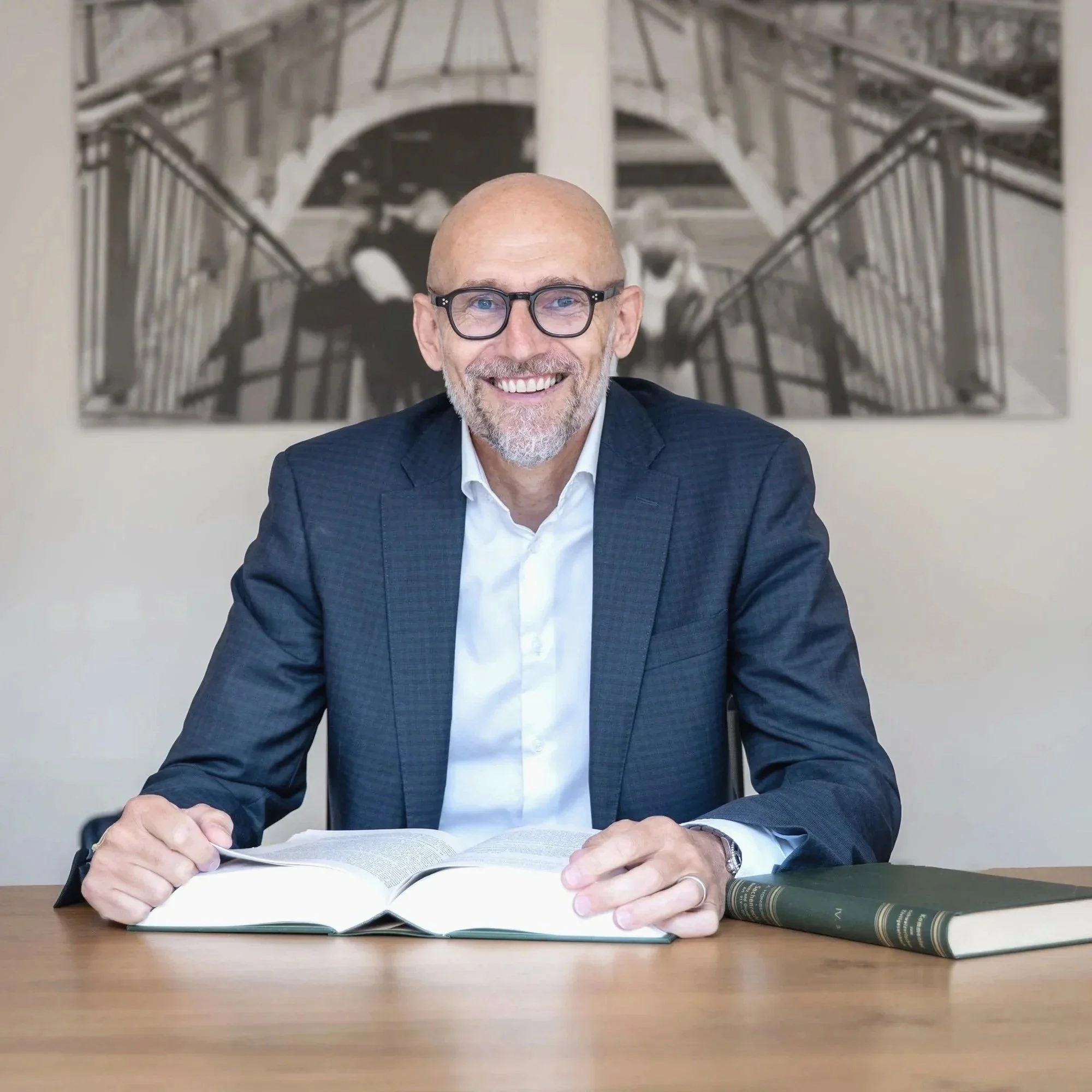 Ein älterer Mann mit Brille und grauem Bart liest in einem Buch an einem Schreibtisch, im Hintergrund ein Schwarz-Weiß-Foto einer Treppe.