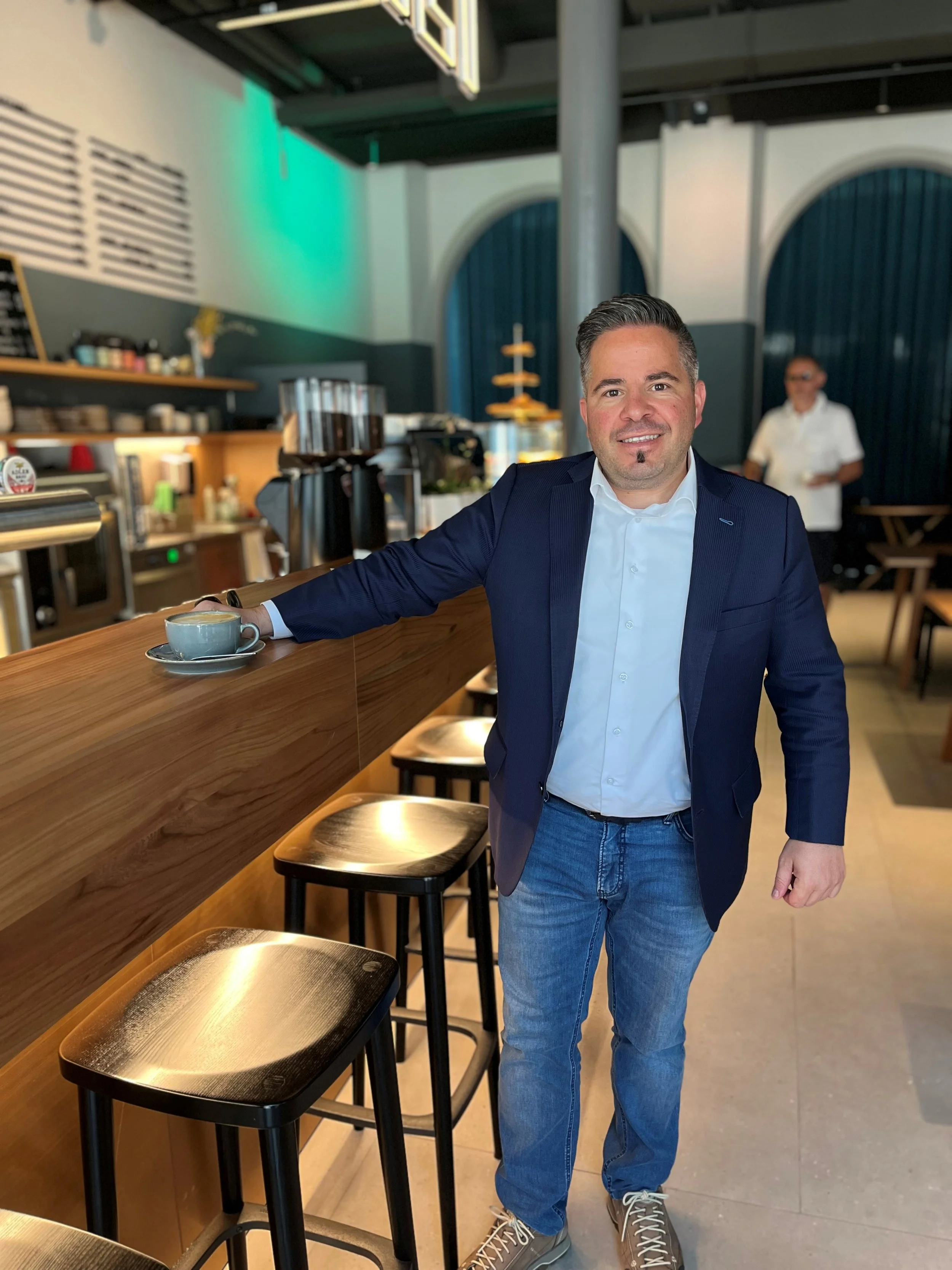 Ein gutaussehender Mann in einem blauen Anzug und blauer Jeans, der in einem Café oder Bistro an einem Holztresen sitzt, zeigt ein freundliches Lächeln. Im Hintergrund sieht man eine Person in einem weißen Hemd und Brille, die in den Raum schaut.