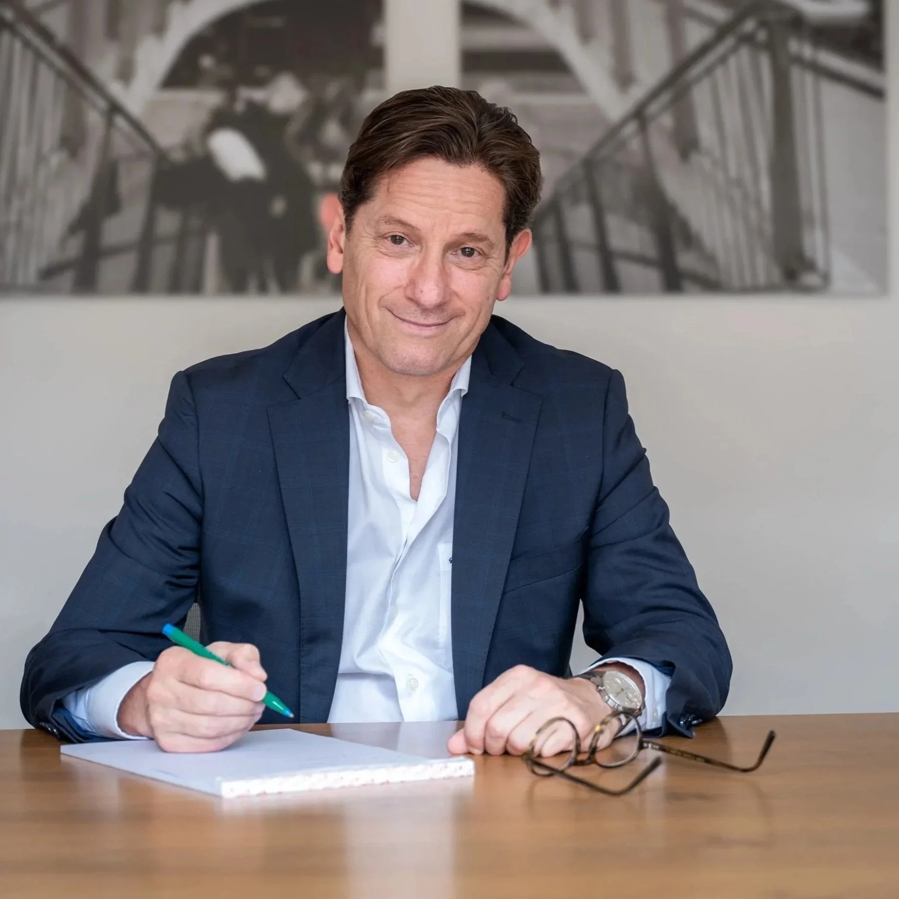 Ein Mann in einem blauen Anzug, der auf einem Holzschreibtisch sitzt, einen Stift in der Hand hält, vor einem weißen Blatt Papier, mit Brille und einer Uhr am Handgelenk, im Hintergrund ein schwarzes und weißes Bild an der Wand.