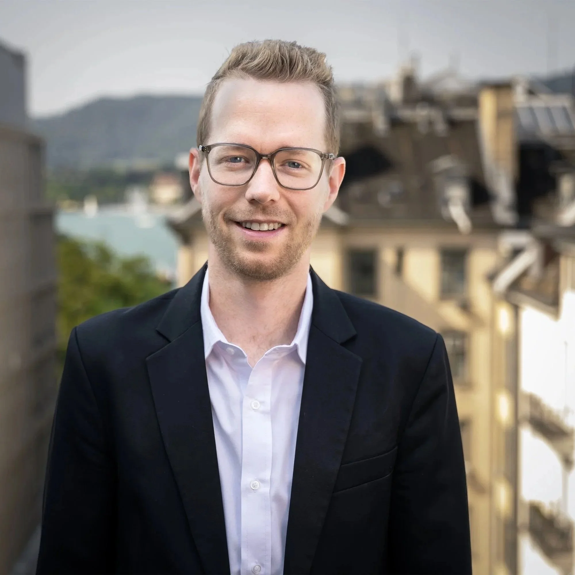 Junger Mann in Anzug und Brille vor einer Brücke mit Wasser im Hintergrund, lächelnd.