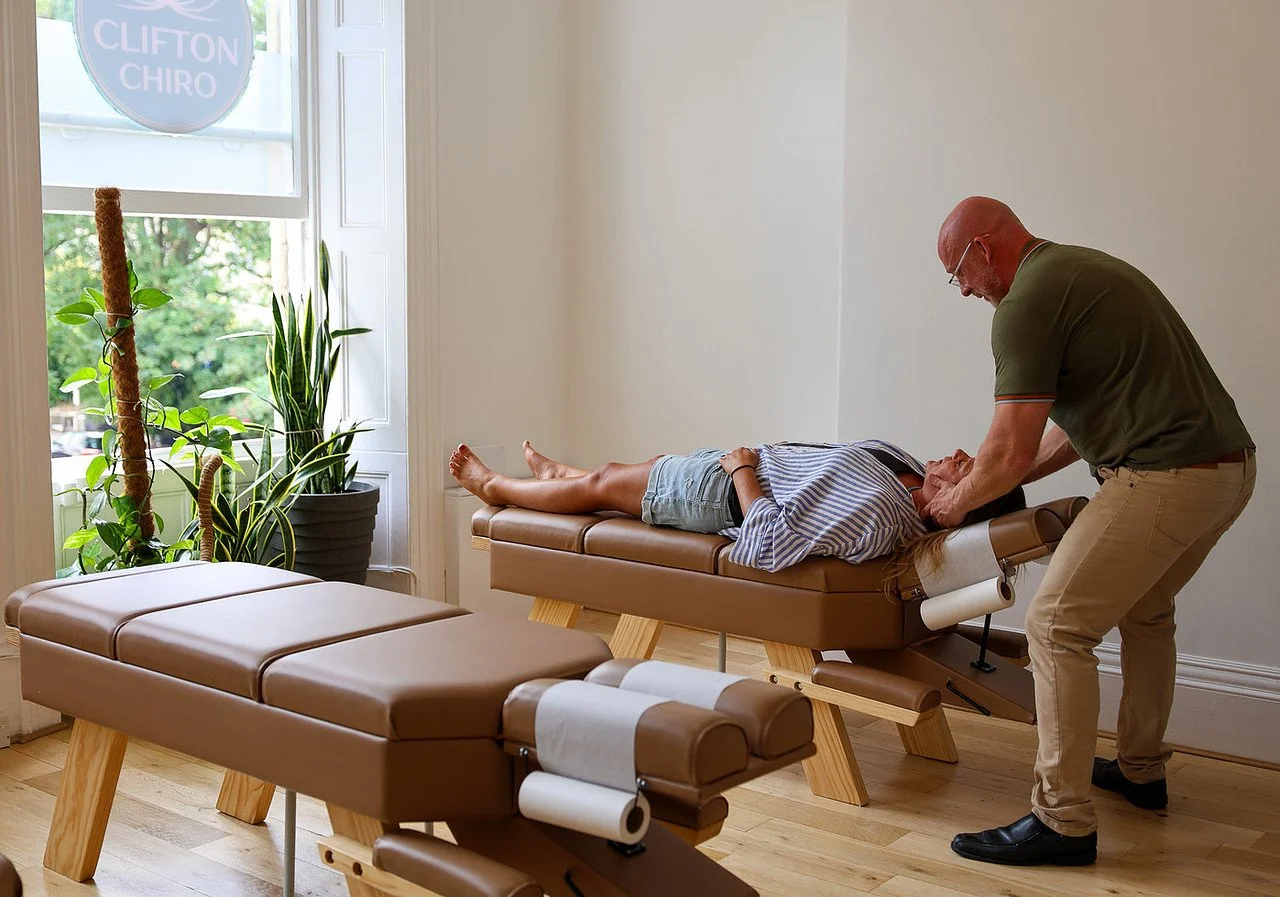 Chiropractor adjusting a patient's neck on a chiropractic table in a well-lit room with plants and a sign that reads 'Clifton Chiro'