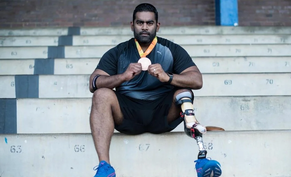 Tyrone a Paralympian with one leg sat on stairs holding onto his gold medal, with a look of pride and dedication on his face.