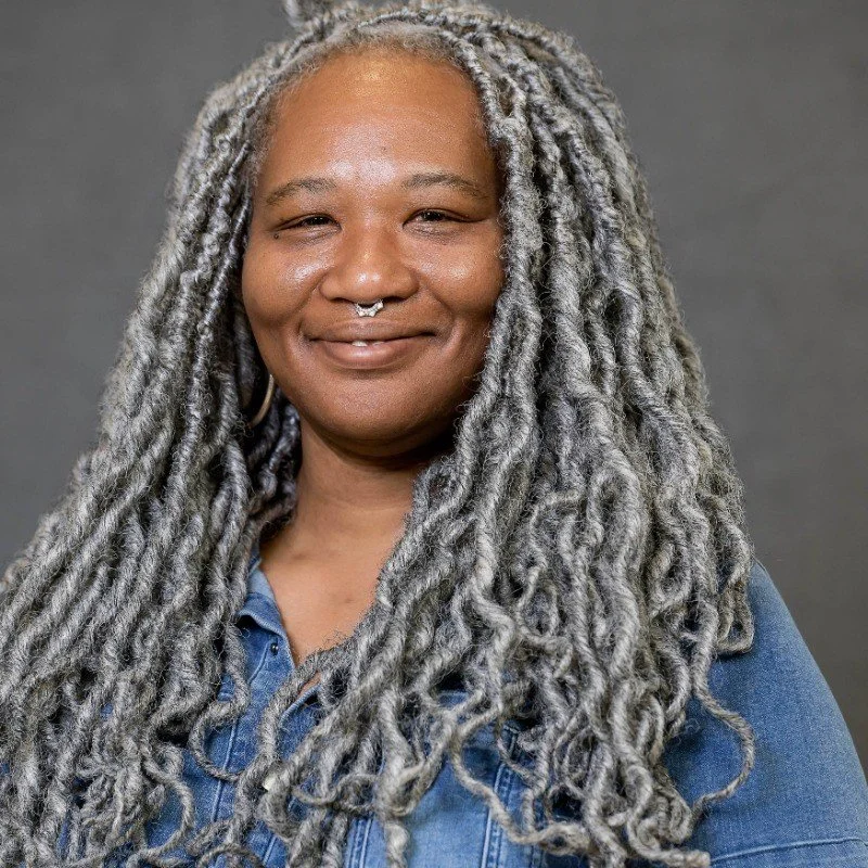 A woman with gray dreadlocks, smiling, wearing a blue denim shirt, with a septum piercing and hoop earrings, against a gray background.