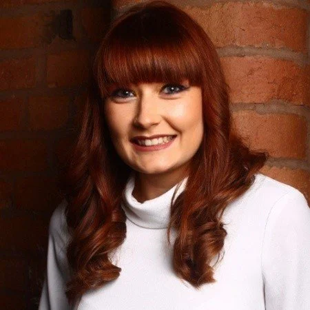 A woman with long, wavy red hair and bangs, smiling, wearing a white turtleneck sweater, standing in front of a brick wall.