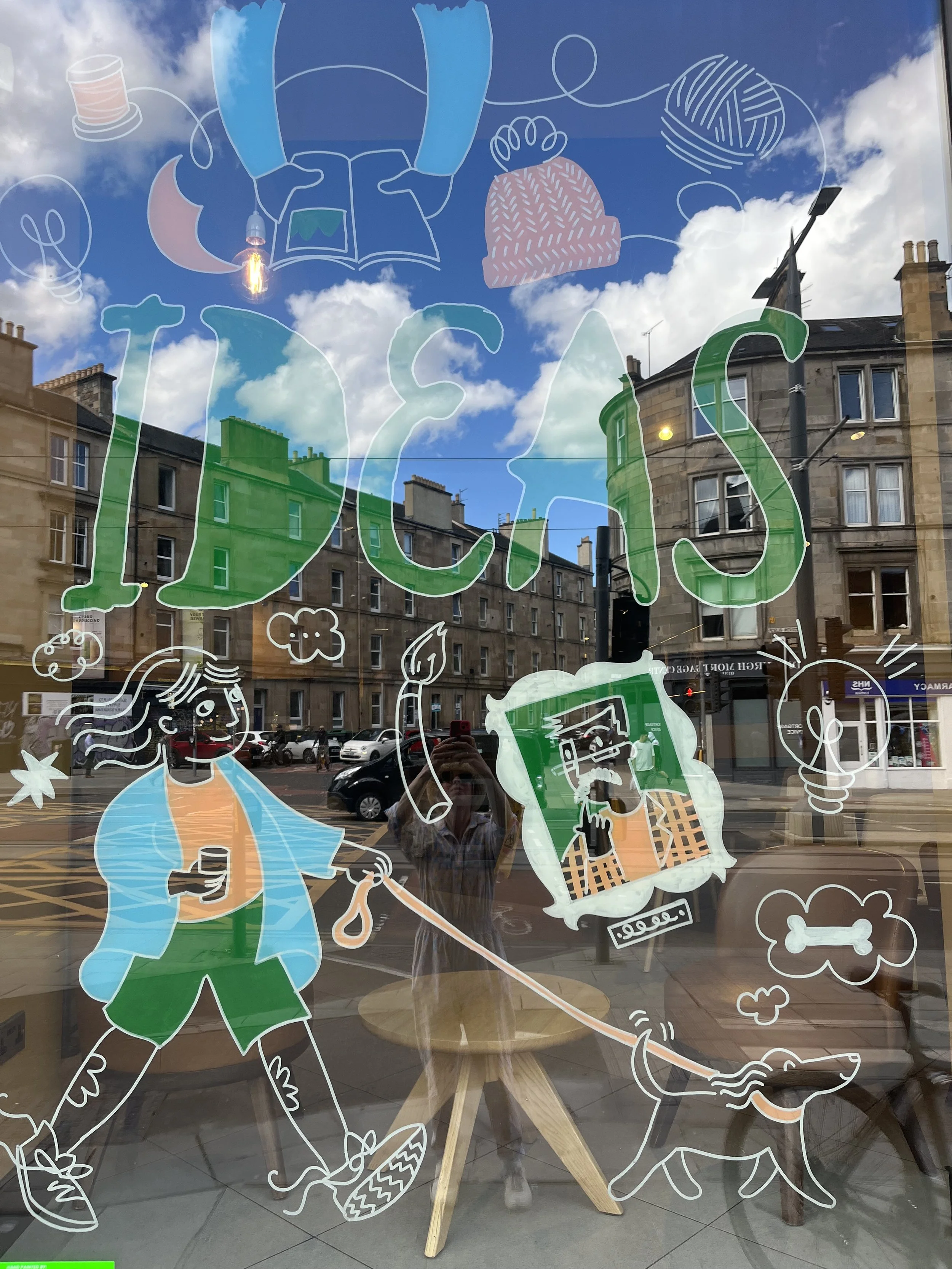 An illustration on a window at Starbucks in Leith Edinburgh walking a dog and taking a coffee on her journey there is also a man in a painting a paint brush with lightbulbs and lettering created by Cassandra Harrison